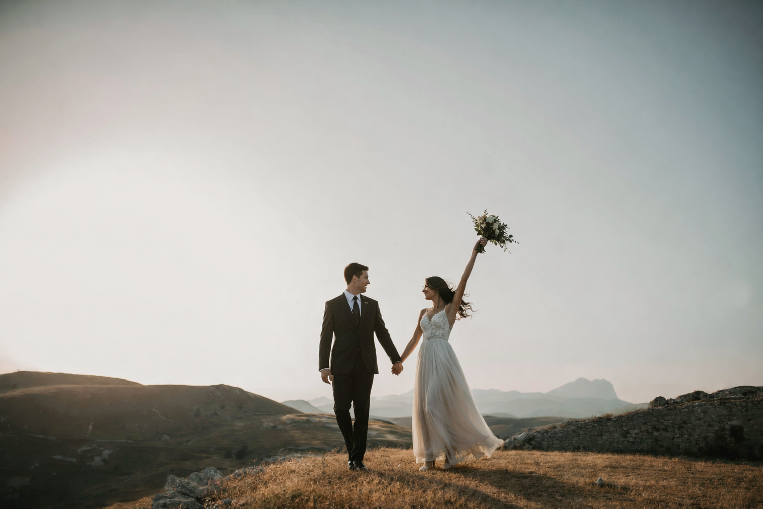 A bride and groom walking hand in hand outdoors on a grassy hill at sunset. The bride is holding a bouquet high in the air and smiling, wearing a white wedding dress, while the groom is dressed in a dark suit, looking at her. The background features 