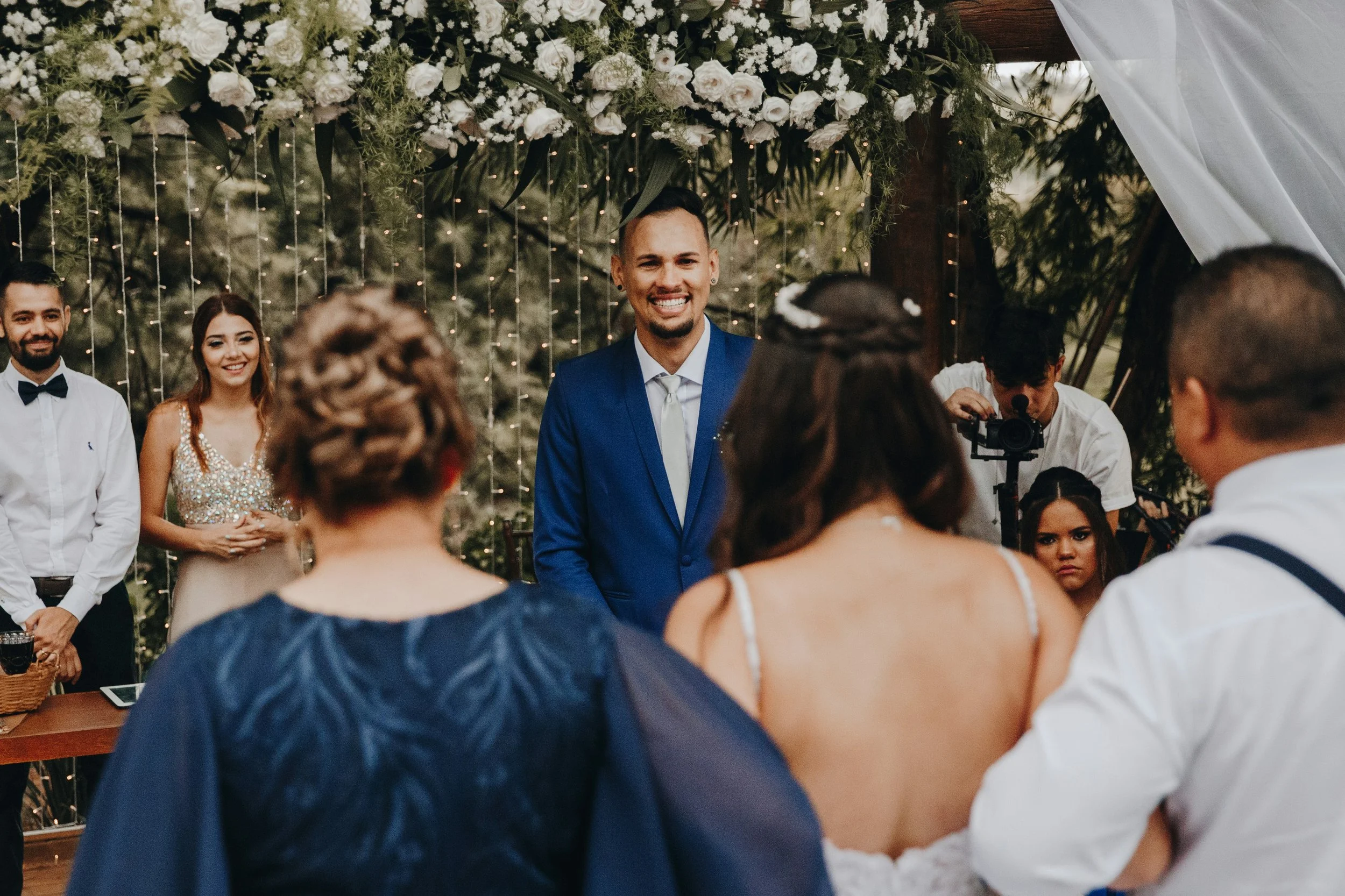 A couple standing together during an outdoor wedding ceremony, facing their celebrant while guests look on.