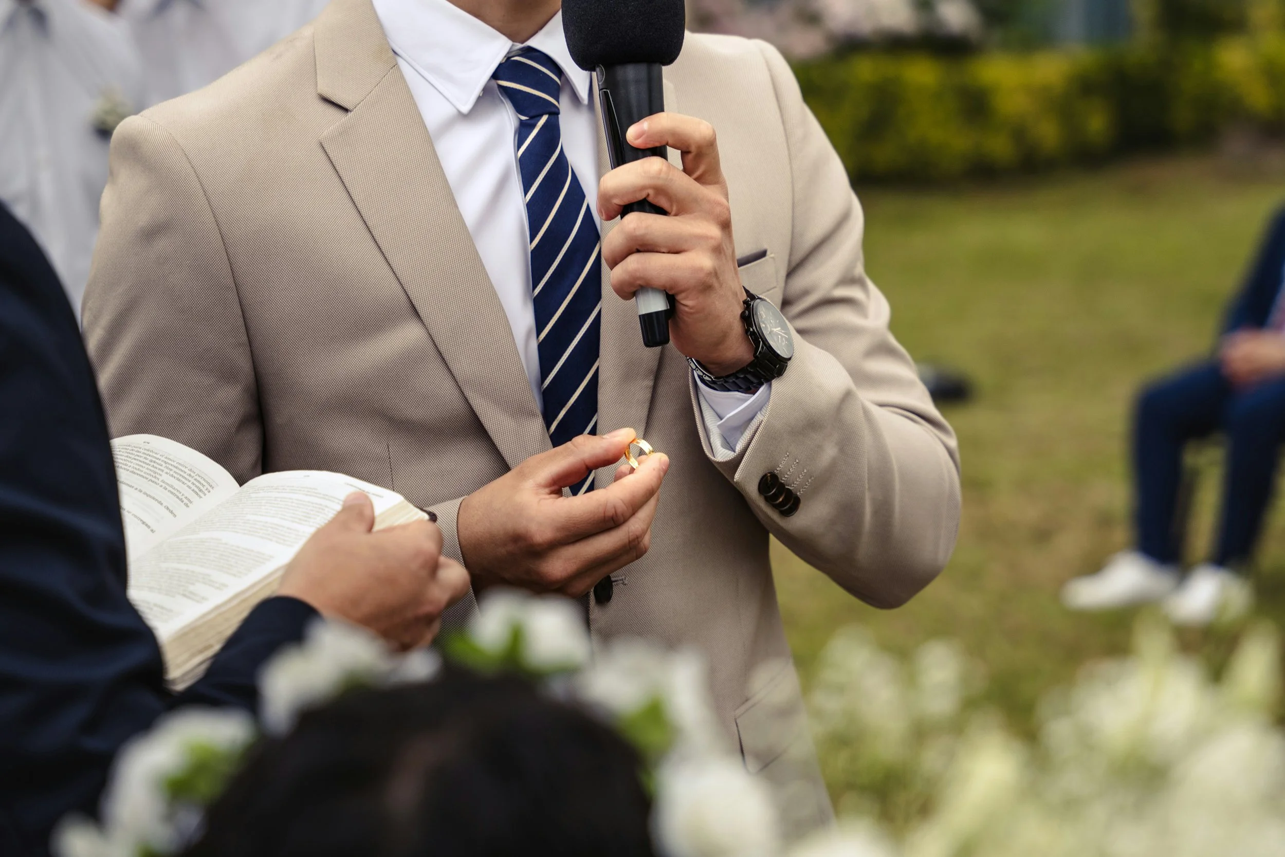 A groom speaking into a microphone during an outdoor wedding ceremony while guests listen.