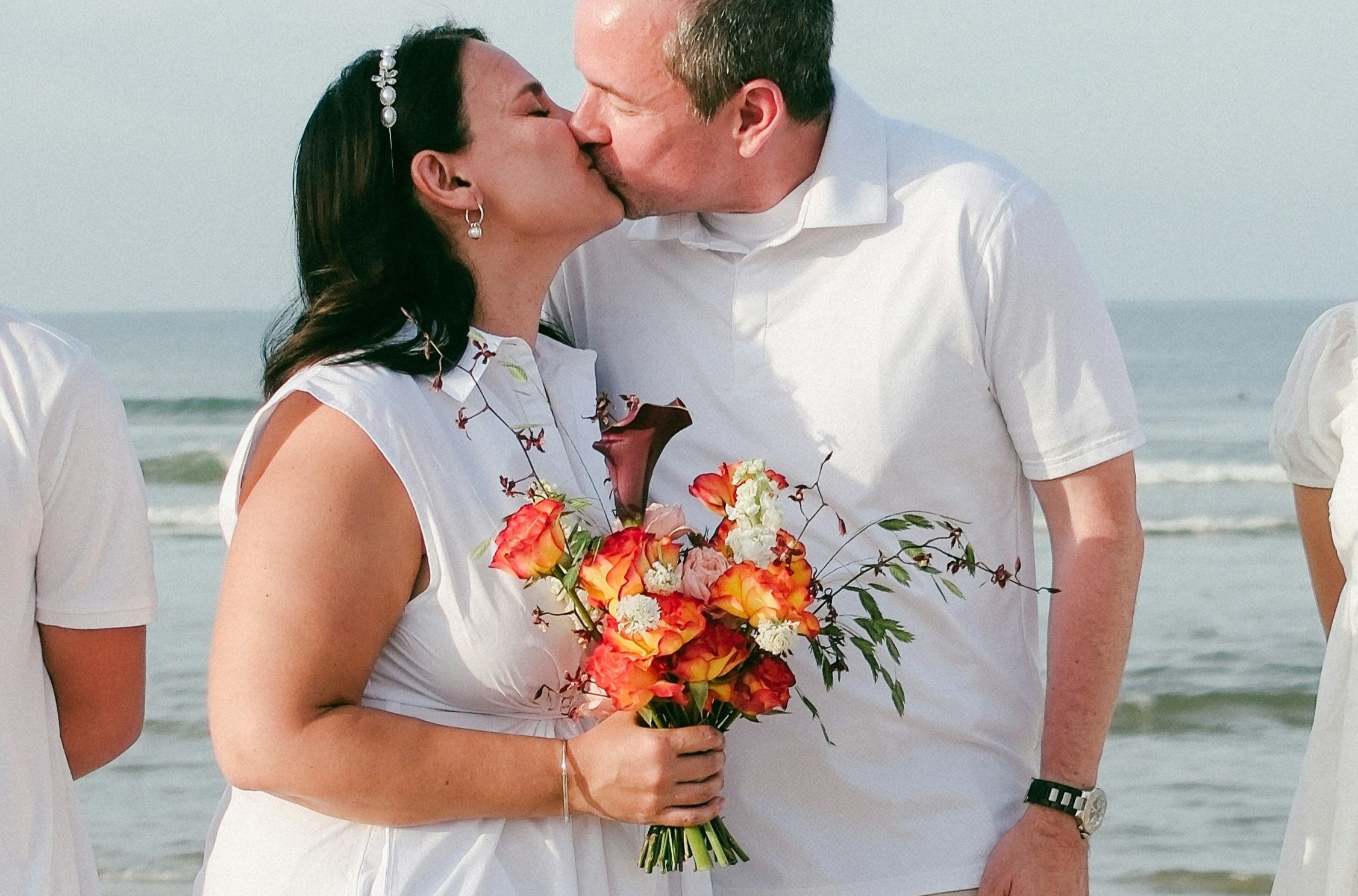 Couple sharing a kiss during a beach vow renewal ceremony, holding a colourful bouquet by the sea.