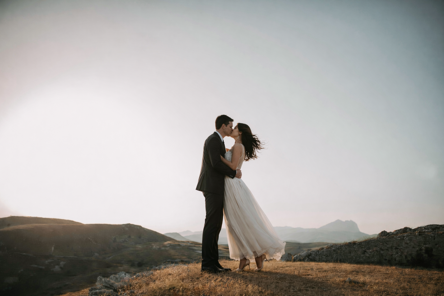 A bride and groom sharing a kiss on a hilltop at sunset, with rolling hills and mountains in the background.