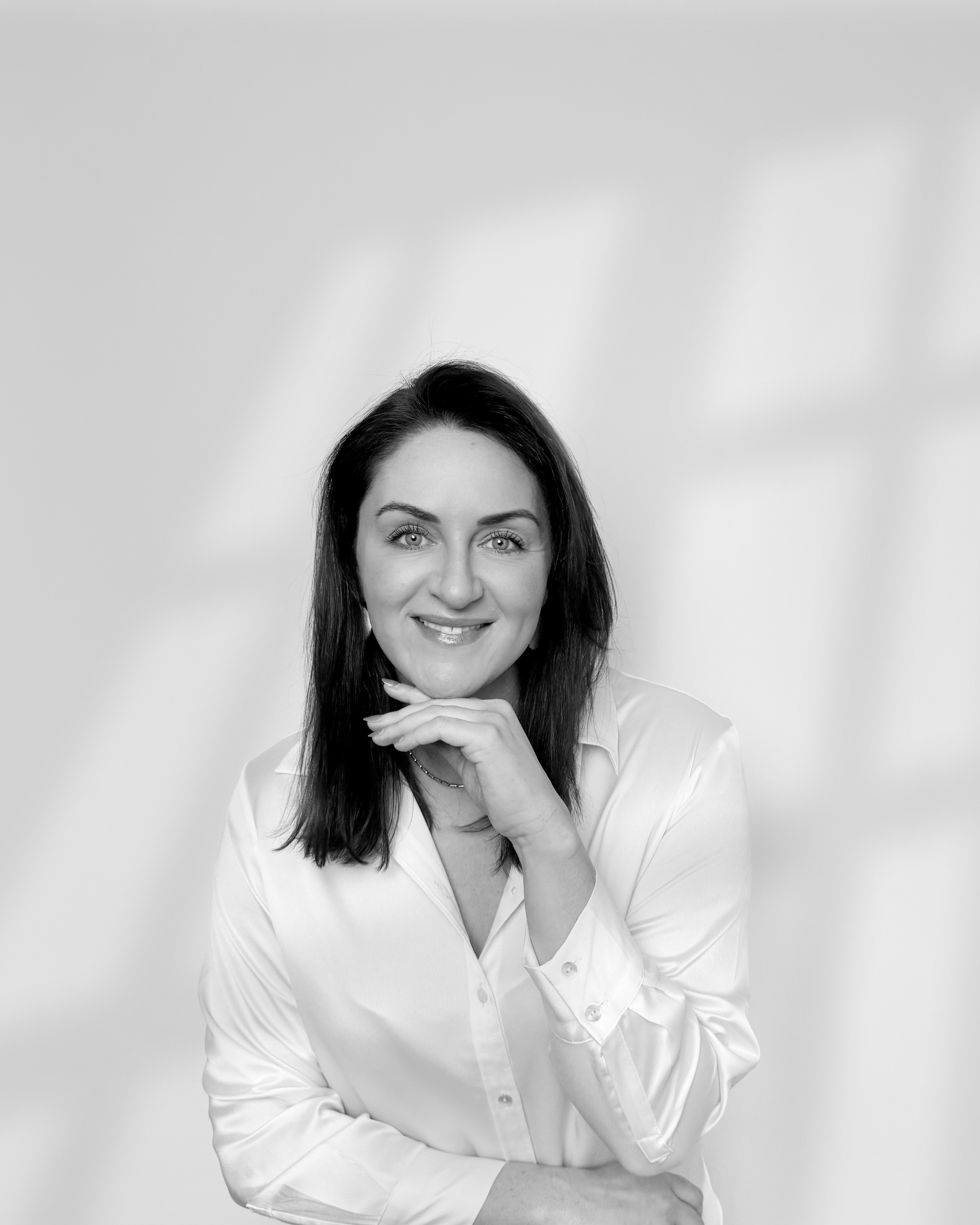 Black and white portrait of Sarah Precious from Sunday Best Ceremonies who has dark brown hair, smiling, wearing a button-up shirt, posing with her hand under her chin against a plain background.