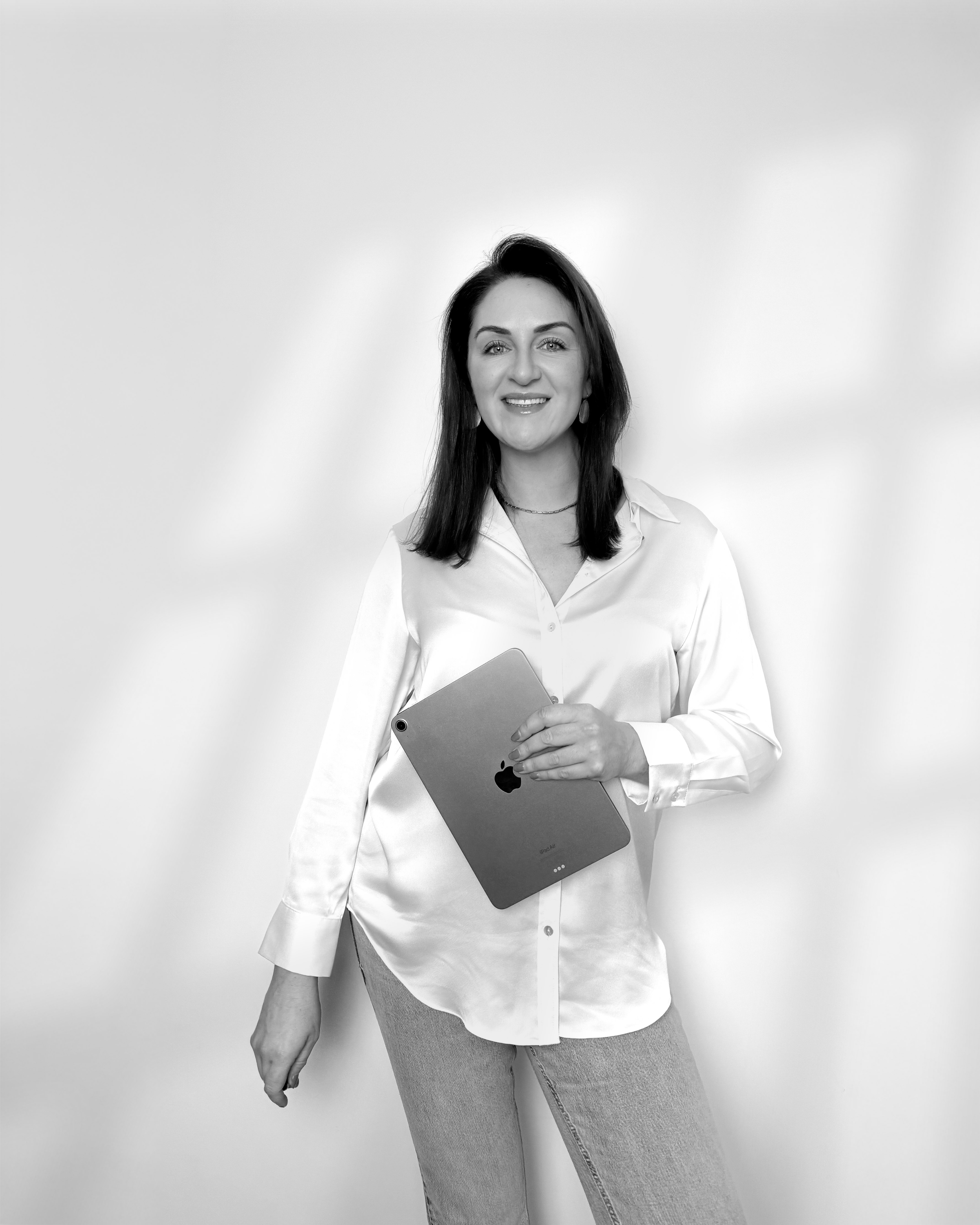 Sarah Precious from Sunday Best Ceremonies who is smiling, wearing a white button-up shirt, holding an iPad, standing against a plain background.