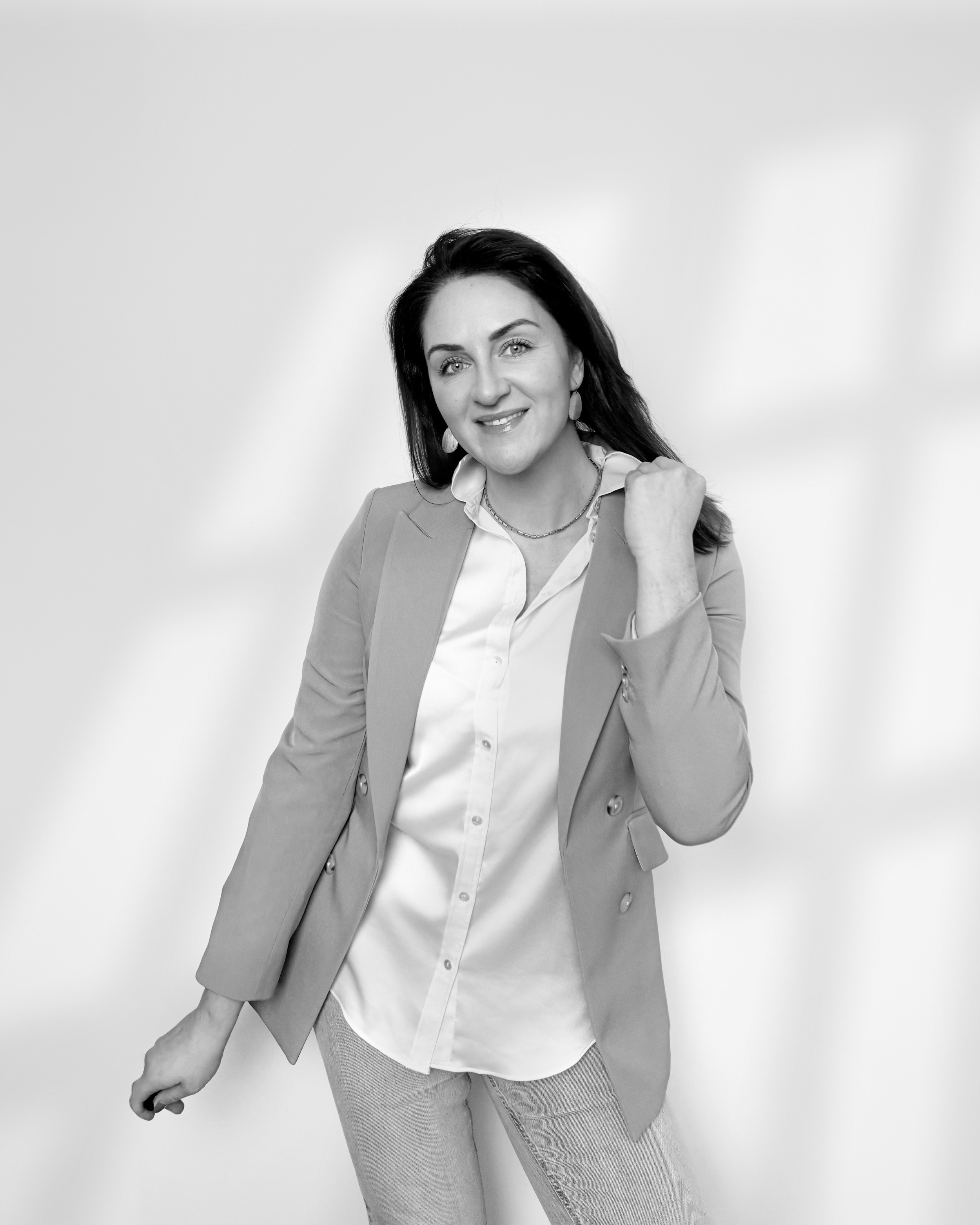 Black and white portrait of Sarah Precious from Sunday Best Ceremonies wearing a light-colored blazer, white shirt, and jeans, smiling and gesturing confidently.