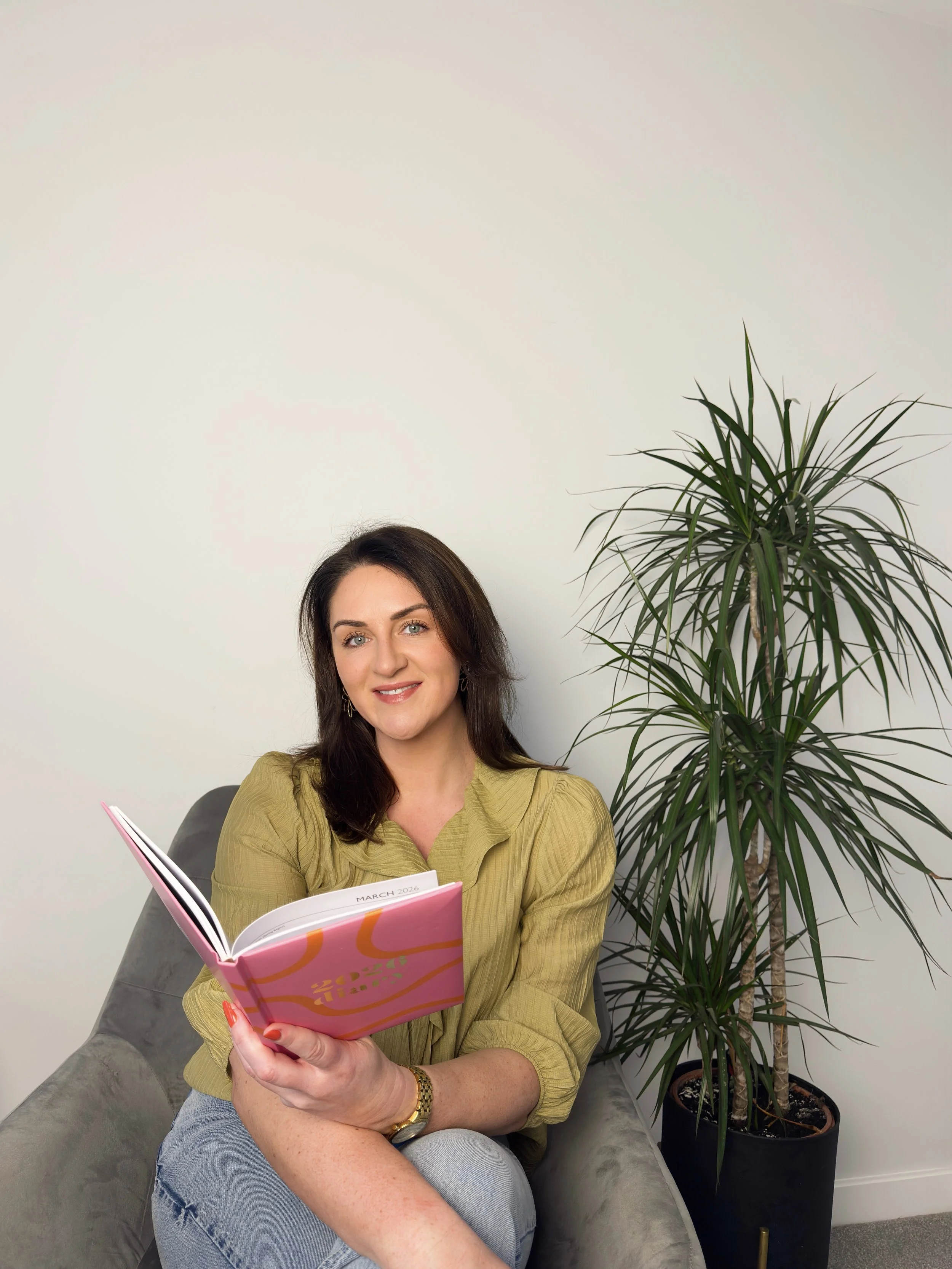 Sarah Precious, celebrant at Sunday Best Ceremonies who has brown hair and blue eyes sitting on a gray sofa, holding a colorful pink and orange planner, smiling, with a large green potted plant beside her against a white wall.