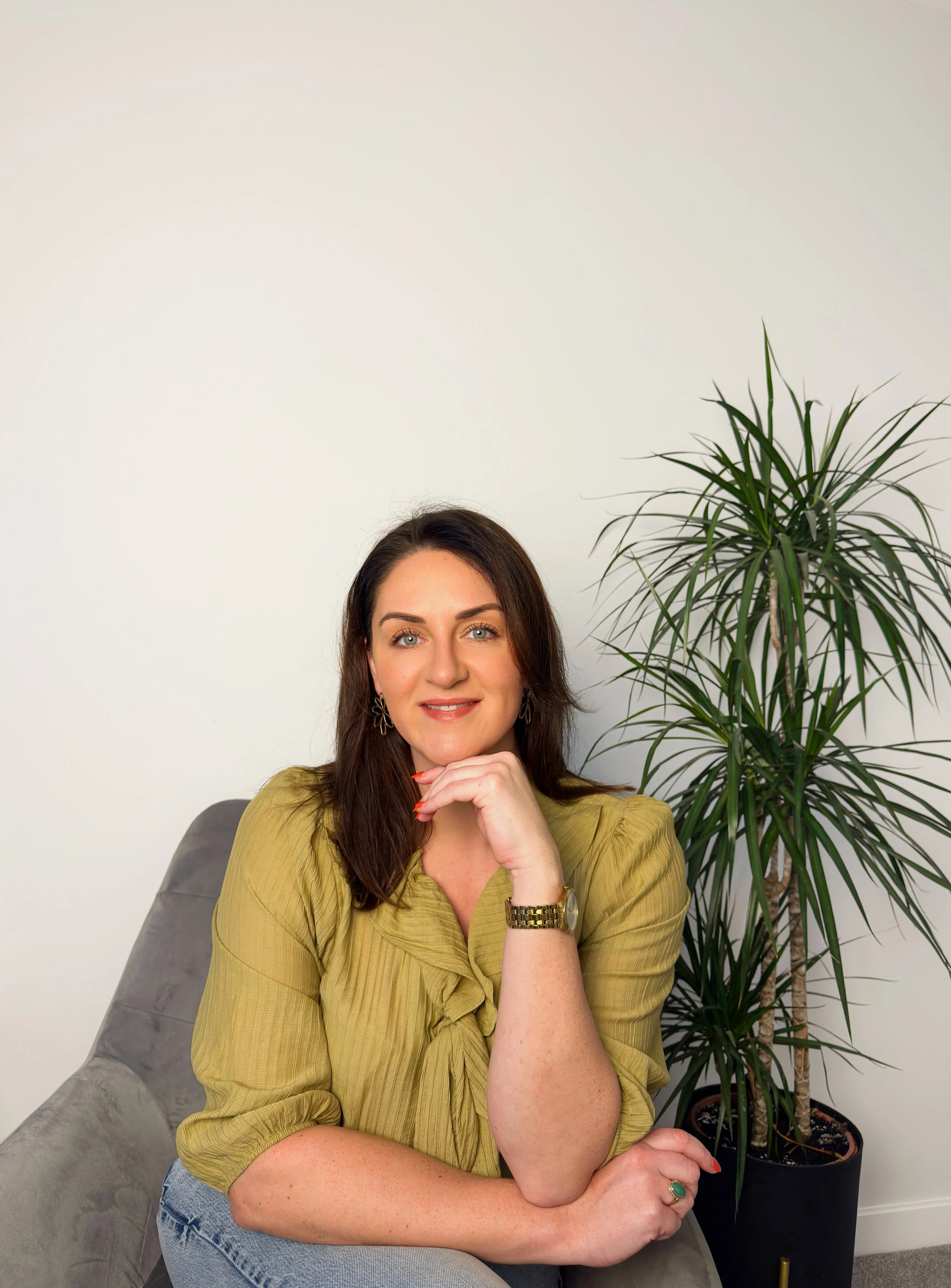 Wedding celebrant Sarah Precious from Sunday Best Ceremonies who has shoulder-length brown hair and green eyes, wearing a mustard yellow blouse, sitting on a grey couch, with a large potted plant in the background.