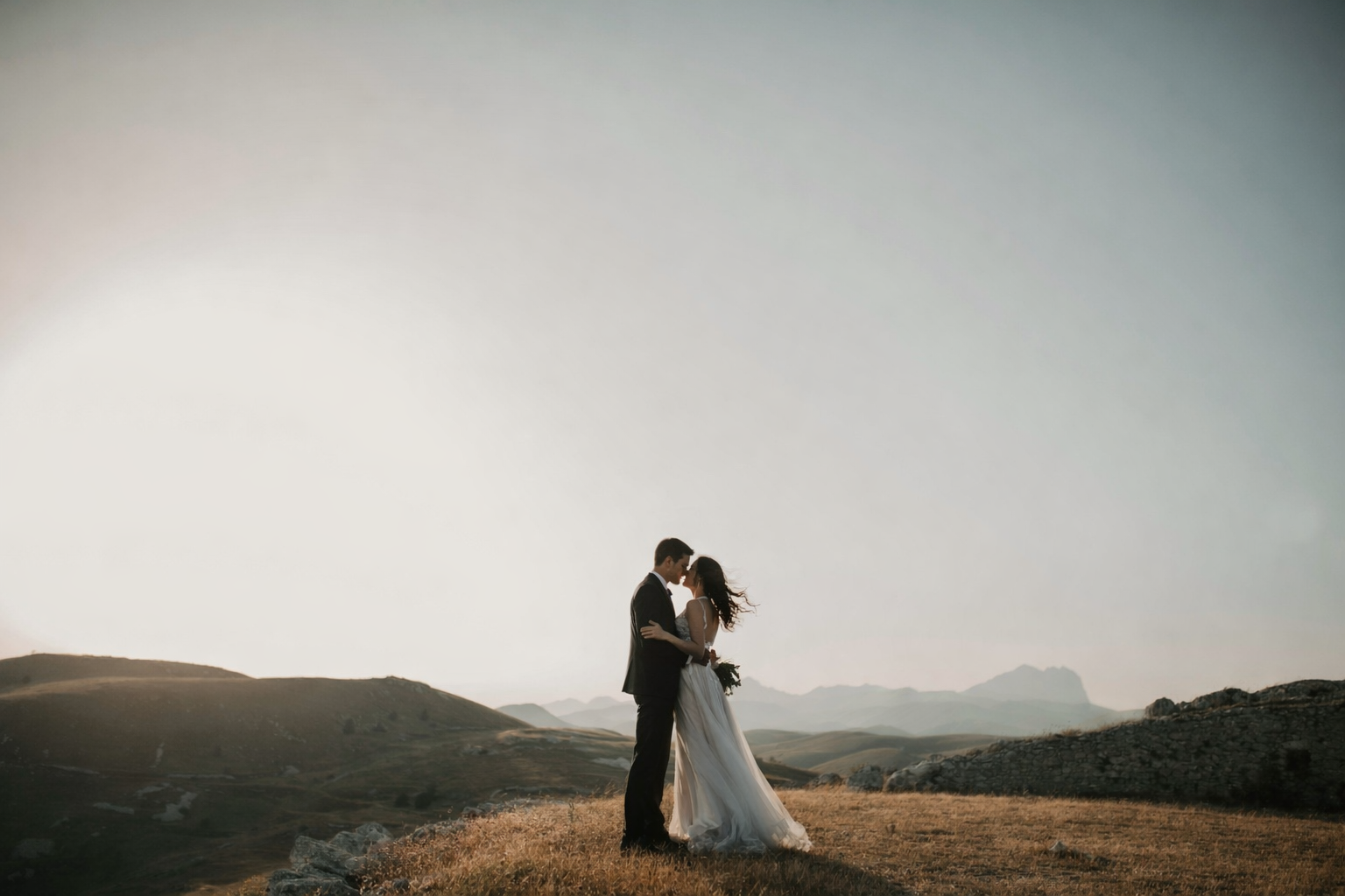 A bride and groom embrace on a grassy hilltop with mountains in the background during sunset.