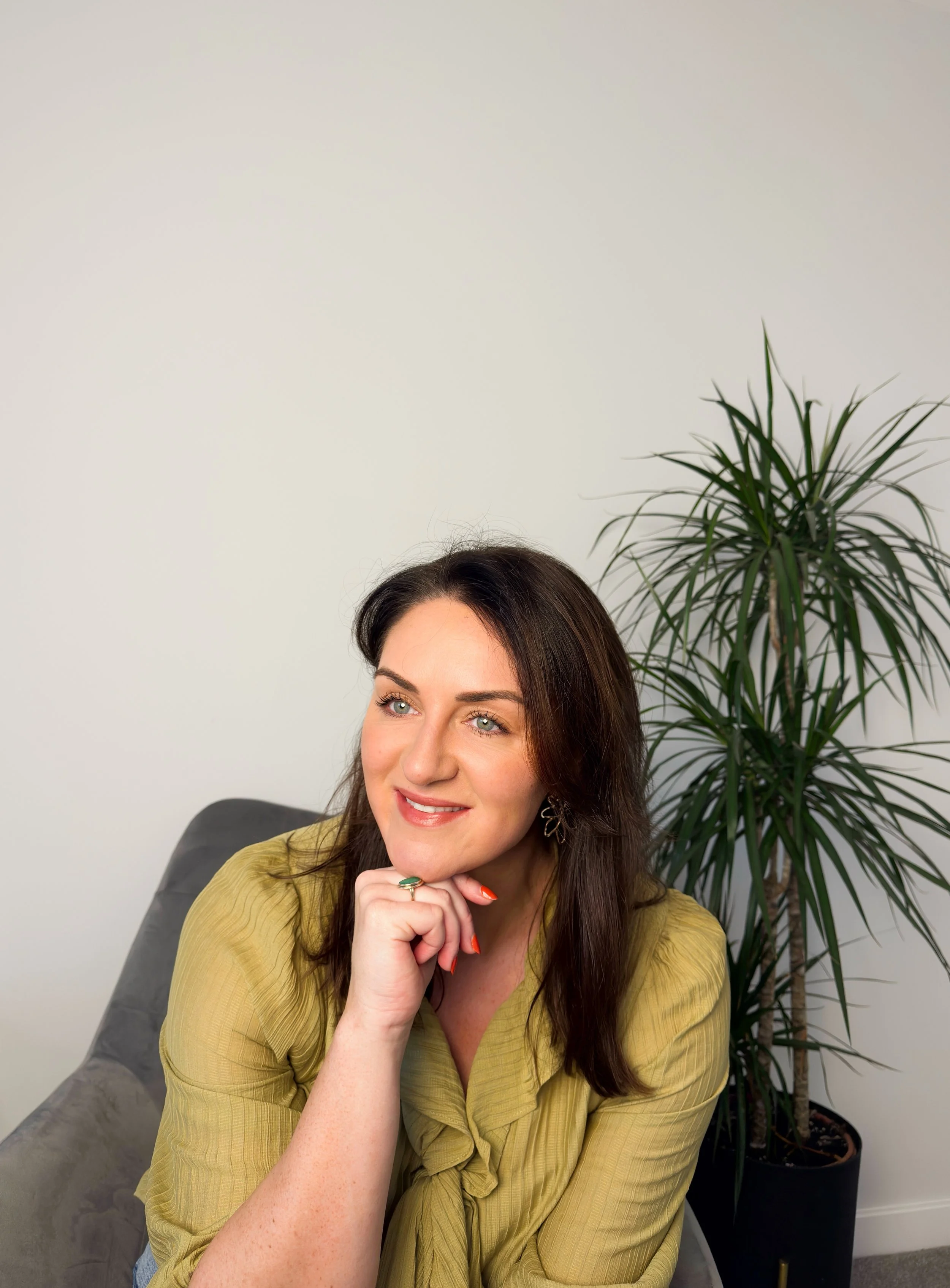 Sarah Precious, the celebrant behind Sunday Best Ceremonies who has long dark brown hair and light skin sitting on a grey couch, wearing a green blouse, smiling and resting her chin on her hand, with a tall potted plant behind her.