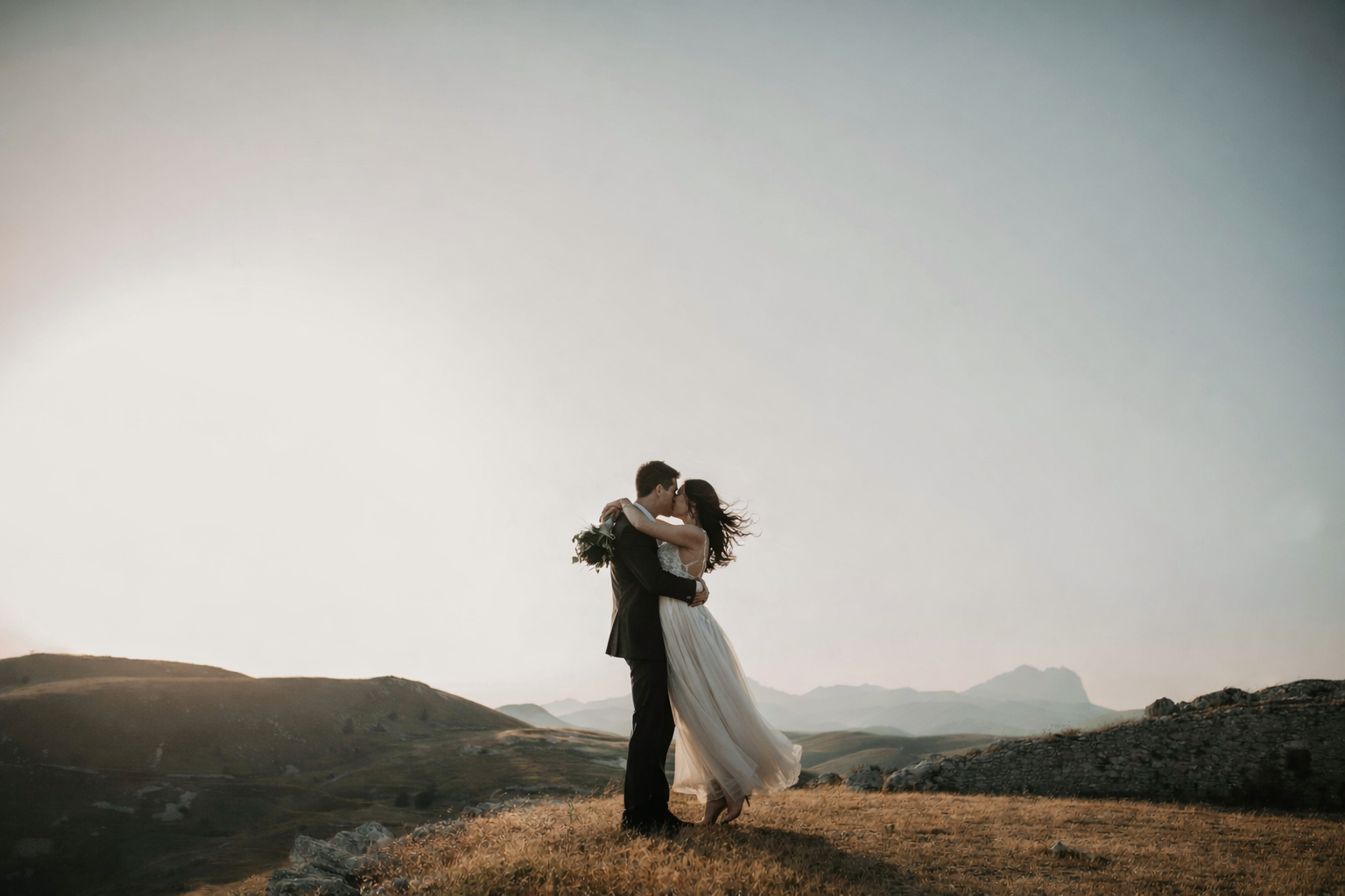 A couple dressed in wedding attire embracing and kissing on a grassy hillside during sunset with mountain range in the background.