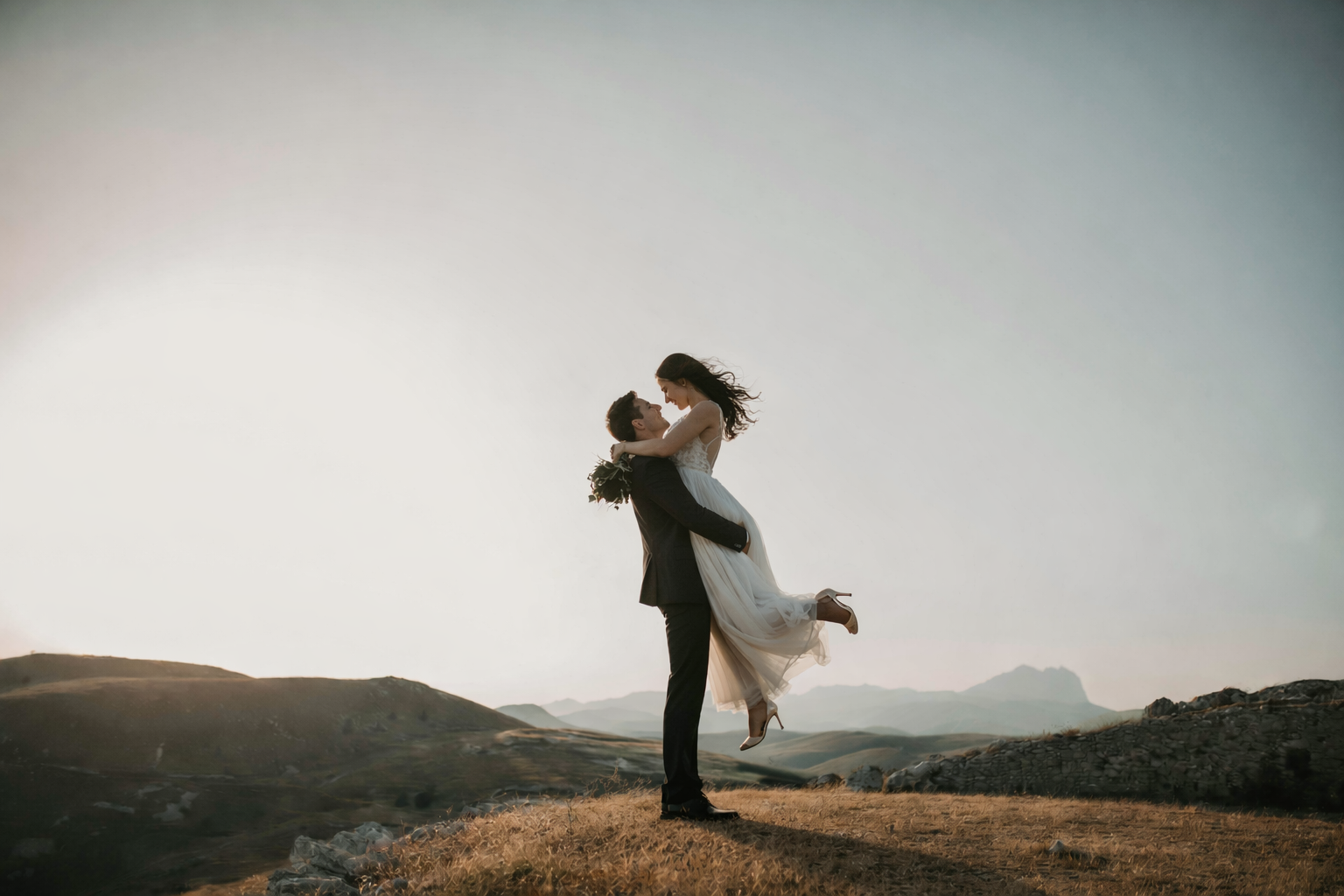 A couple in wedding attire standing on a hill in a vast landscape, with the groom lifting the bride in his arms as they smile at each other against a clear sky.