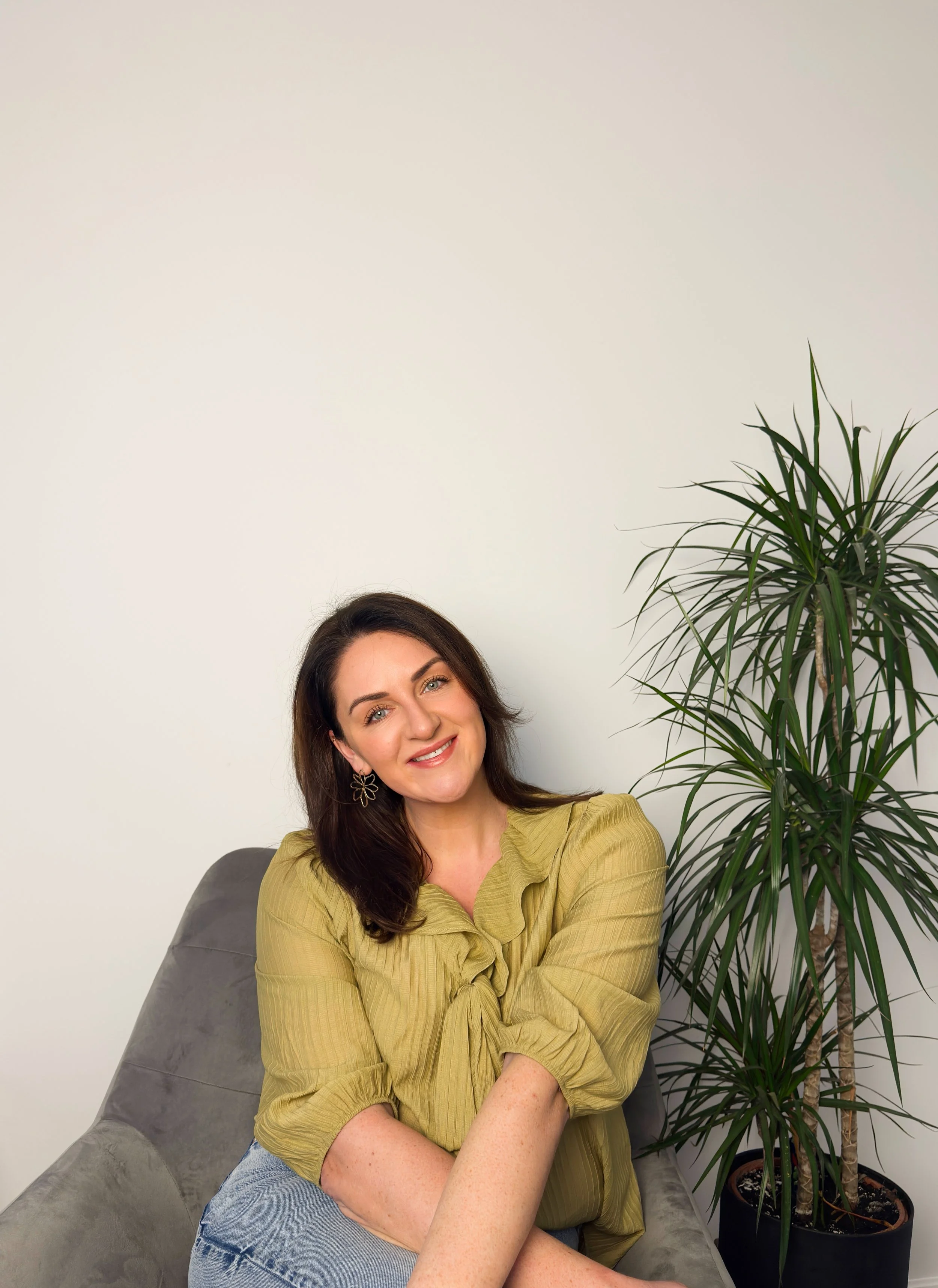 Wedding celebrant Sarah Precious from Sunday Best Ceremonies who is smiling and has dark brown hair sitting on a grey couch, wearing a green blouse and light blue jeans, with a large green potted plant beside her against a white wall.