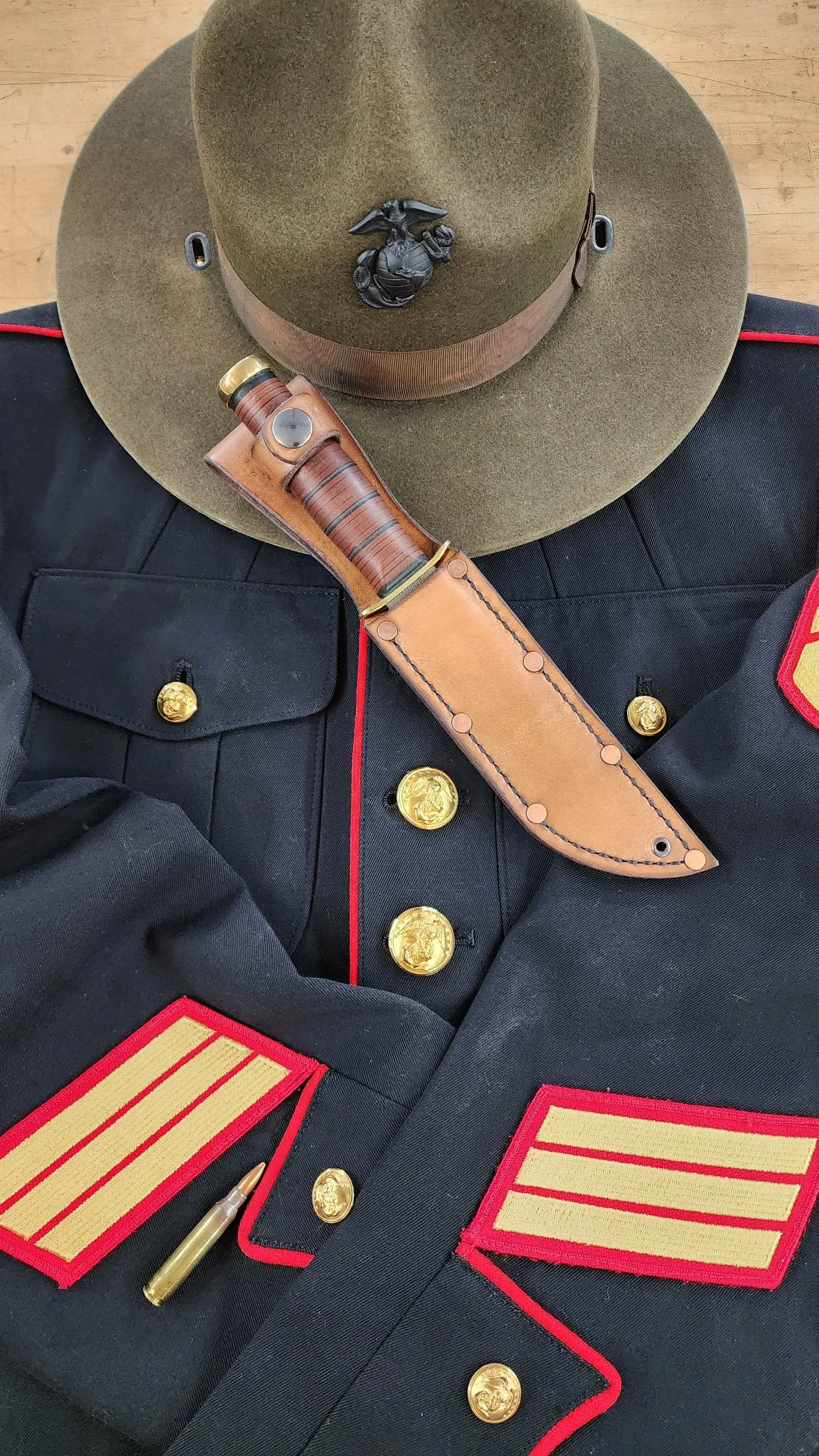 A military dress uniform with gold buttons, red and gold rank insignia, a green hat with a drill badge, a leather knife sheath with a knife, and a bullet.