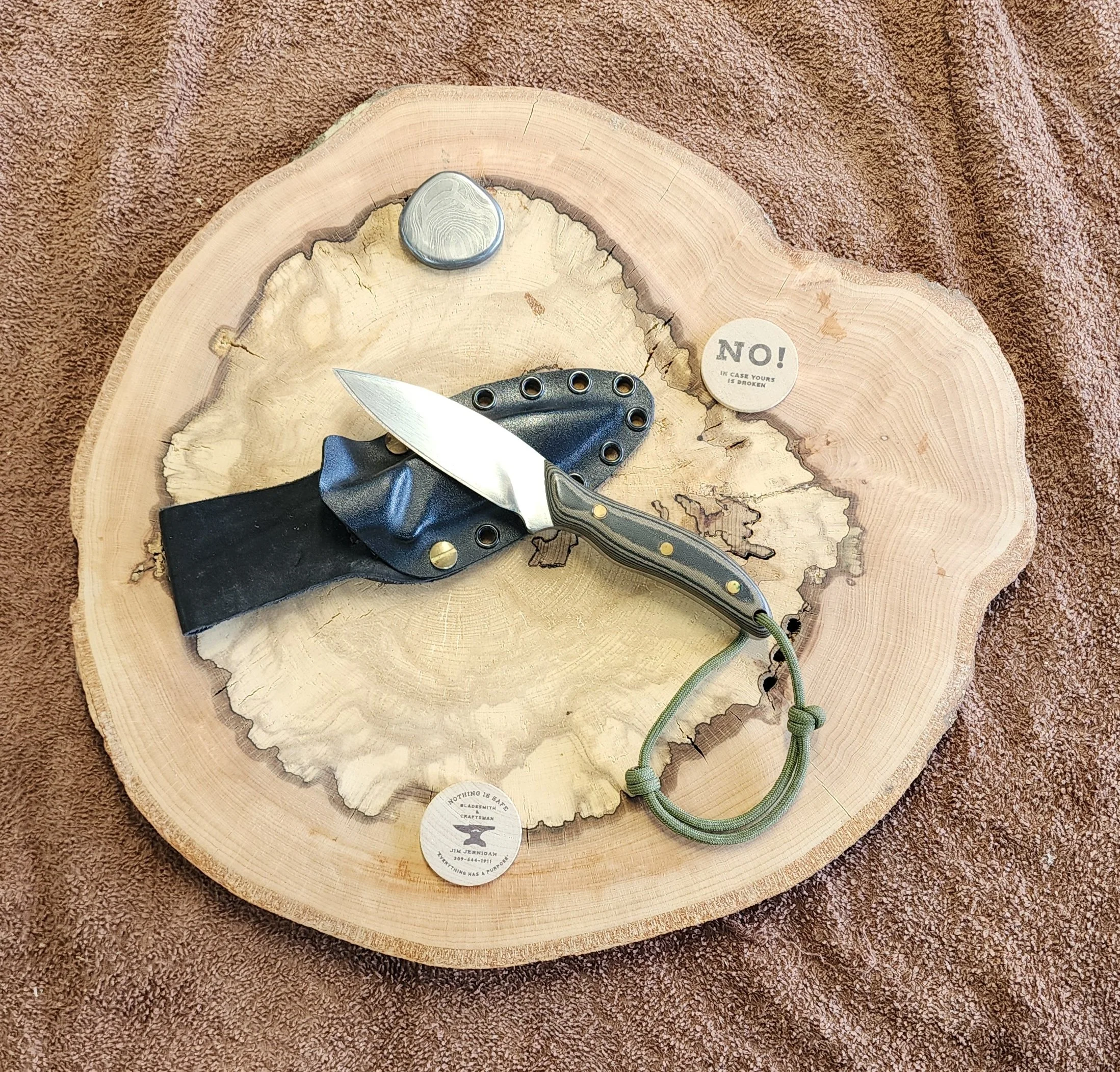 A small fixed-blade knife with a black handle, a black leather sheath with brass rivets, and a round metal object on a wooden slice with bark, placed on a brown fabric background.