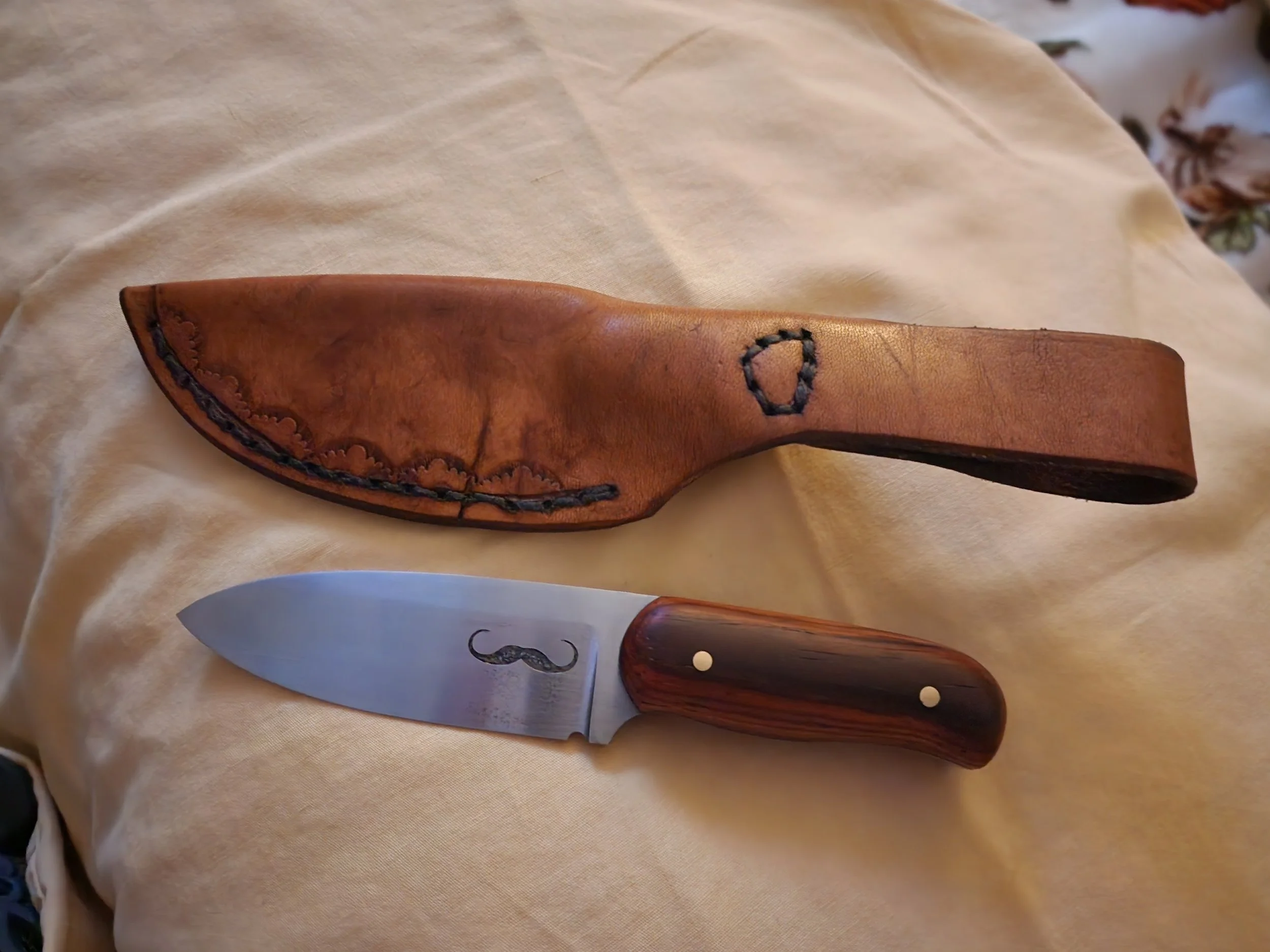 A pocket knife with a wooden handle and a curved blade, placed on a beige cloth. The knife has a metal rivet on the handle and a design on the blade. There is a brown leather sheath with black stitching and a hole for attachment.