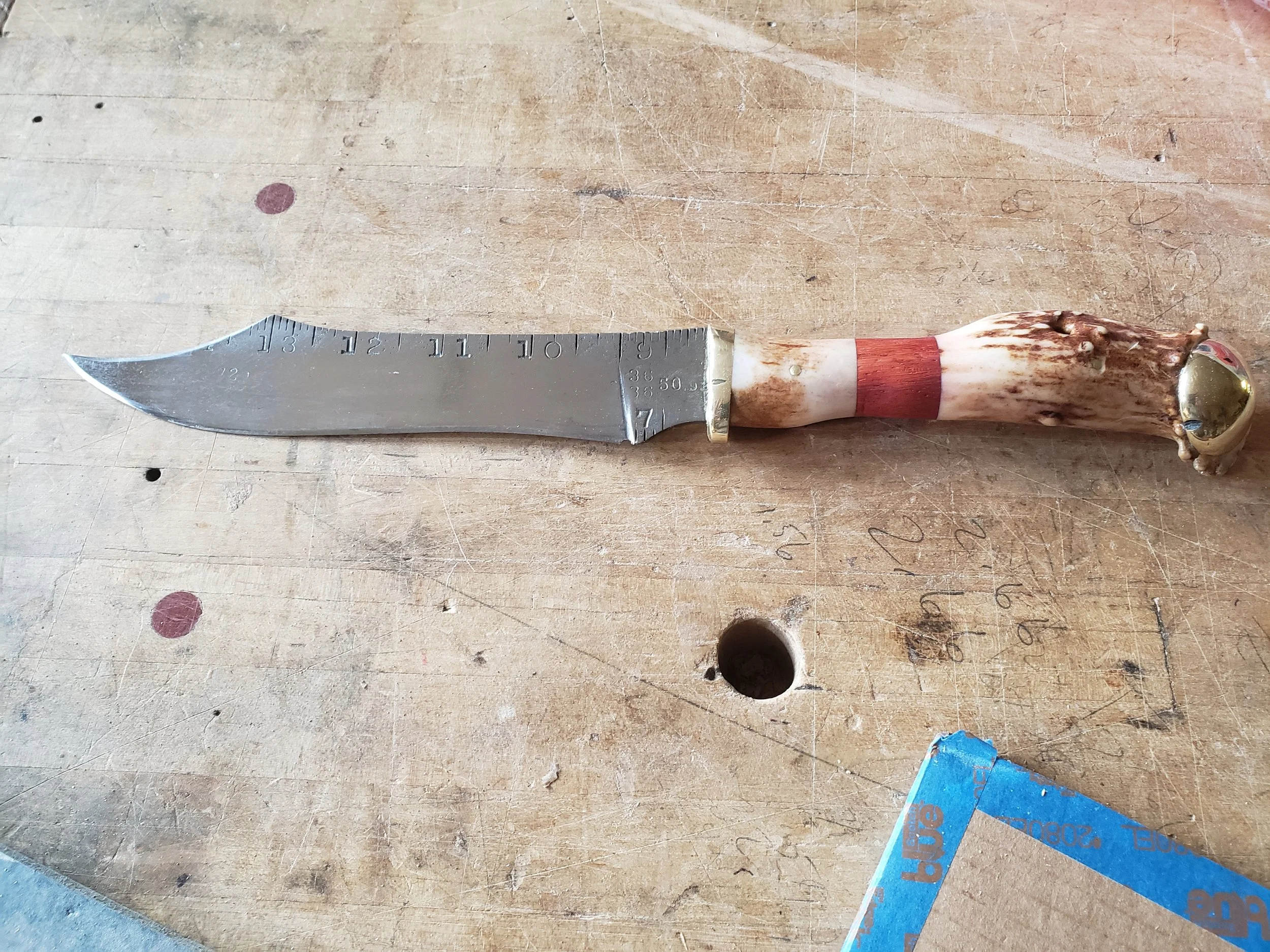 A knife with a blade resembling a measuring ruler and a handle made from antler, placed on a wooden surface with markings and a hole.