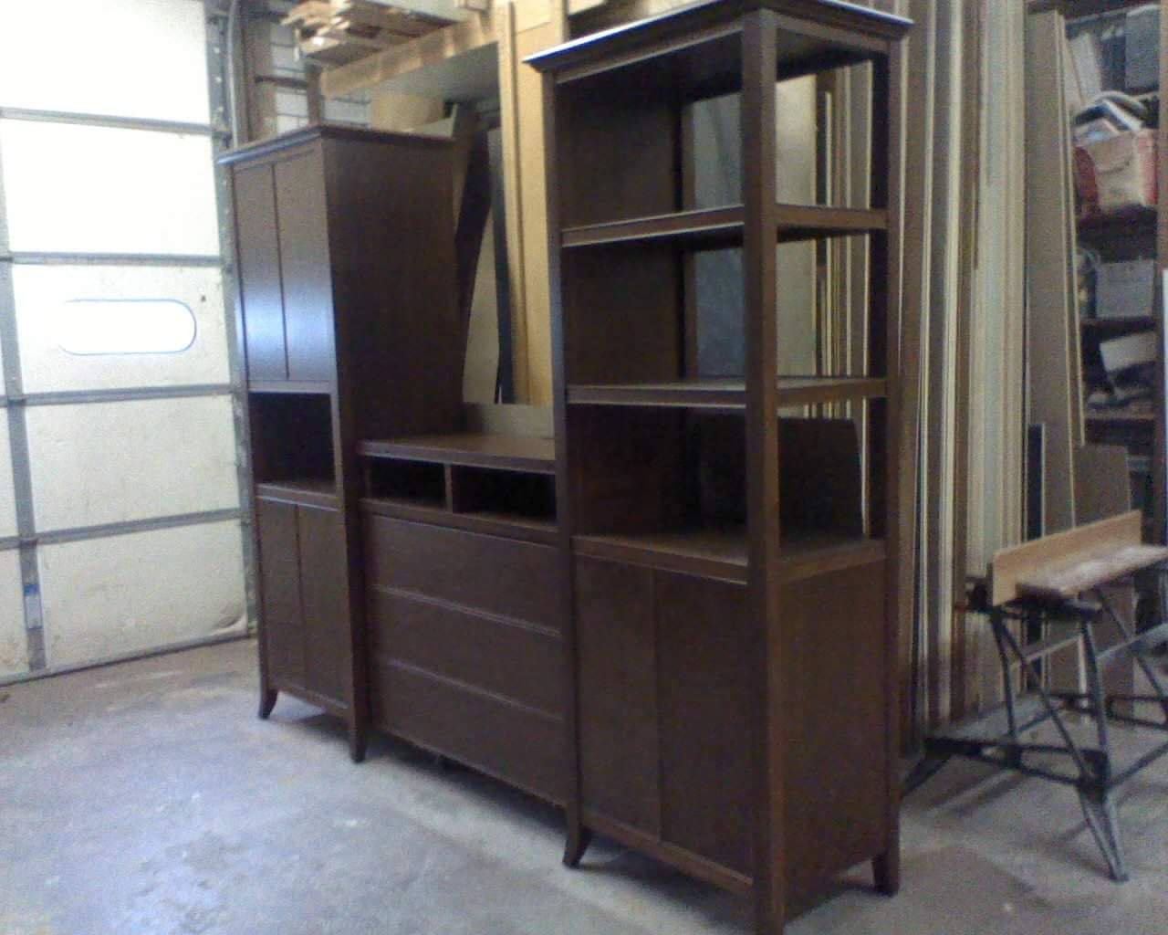 Wooden furniture pieces, including a shelving unit and cabinets, are stored in a garage or workshop space with concrete flooring and a partially visible garage door on the left.