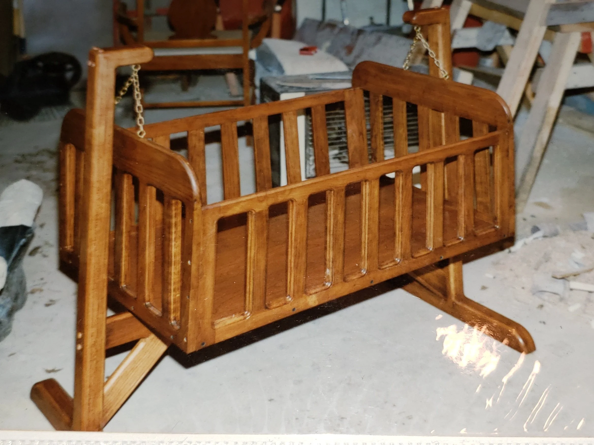 A small wooden rocking cradle with slatted sides and a polished finish, placed in a workshop or storage area.