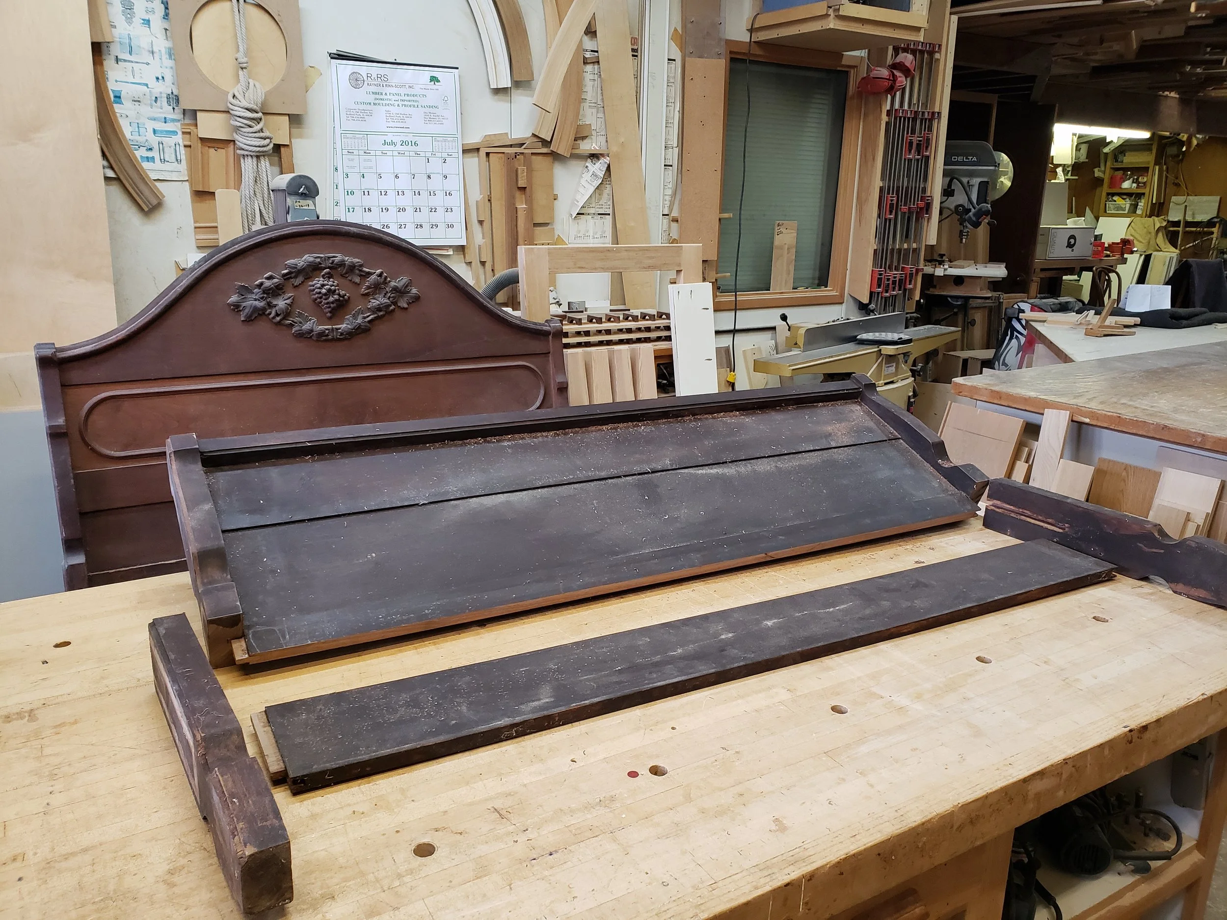 A woodworking workshop with a wooden headboard featuring a carved grapevine design, along with various woodworking tools and materials, including a large wooden tray and pieces of wood on a workbench.