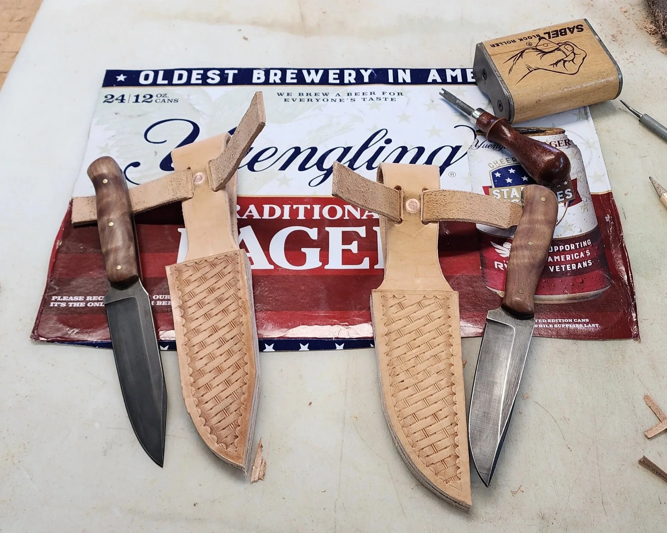 Two partially carved leather and wood-handled knives on a worktable, with a cardboard box and a poster promoting a beer brand in the background.