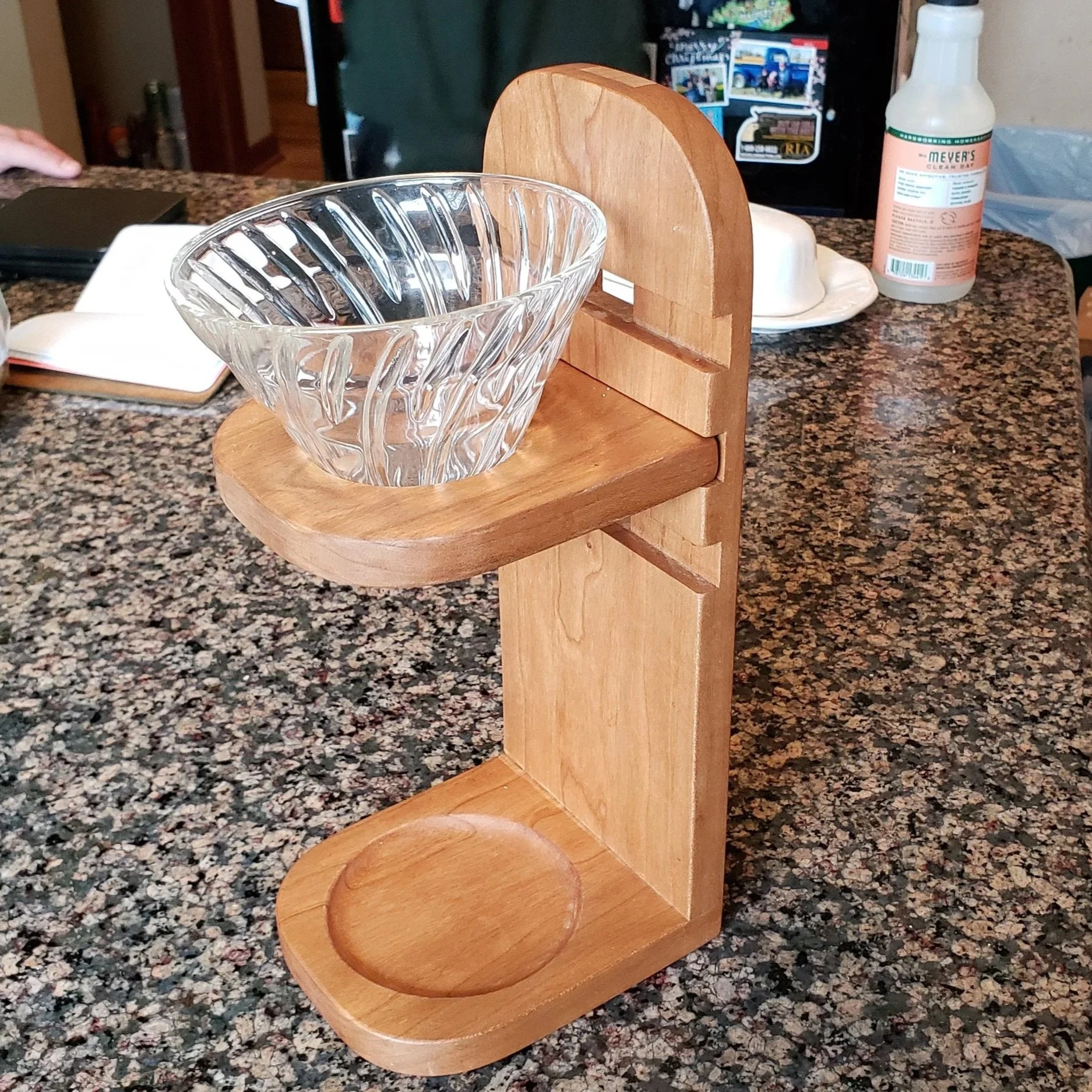 A wooden stand with a glass bowl on the top shelf, placed on a granite kitchen countertop. In the background, there are various kitchen items including a folded notebook, a bottle of cleaning spray, and some plates.