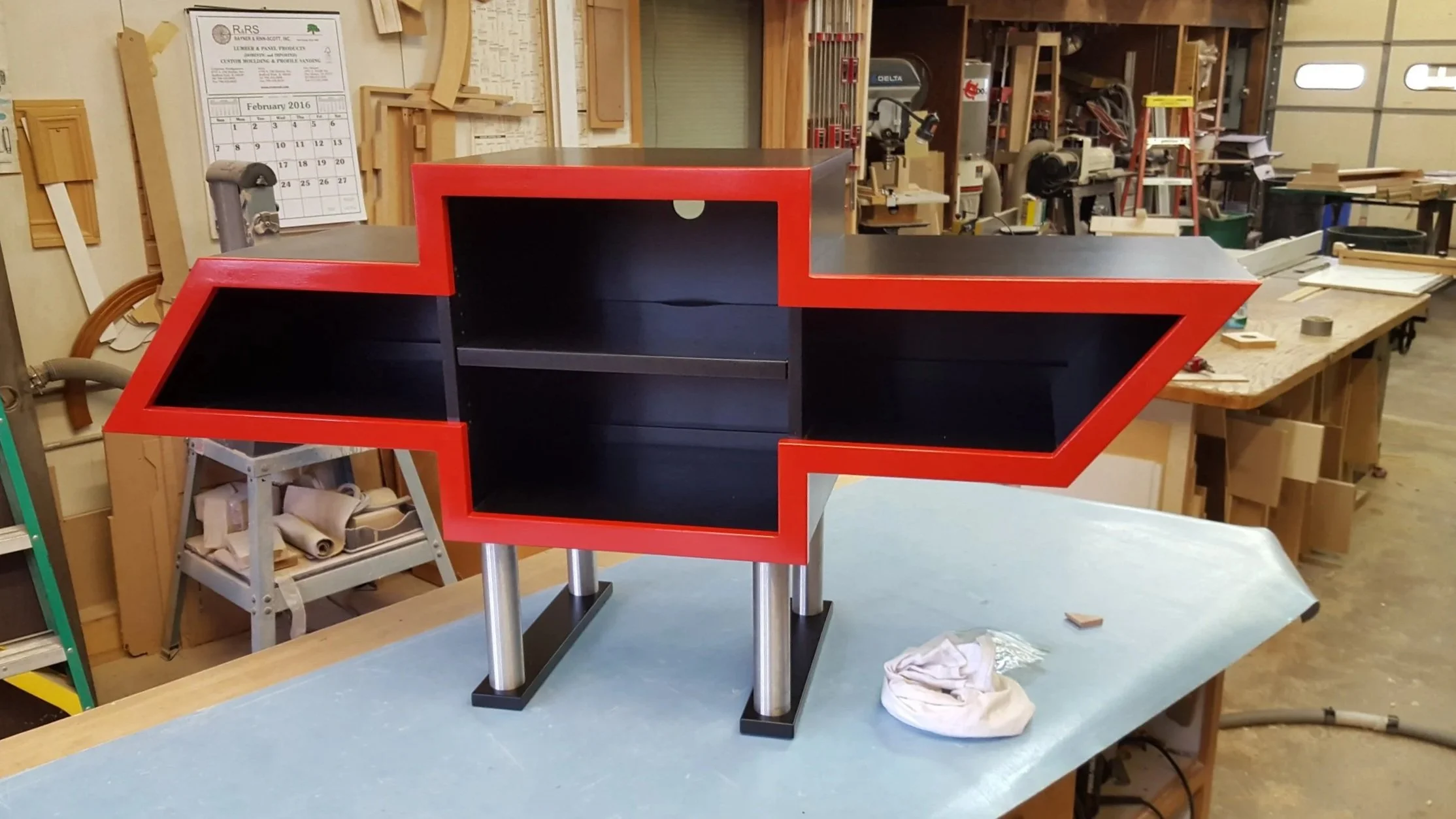 A custom arrow-shaped shelf with a black interior and red outline on a workshop table, supported by four metal legs.