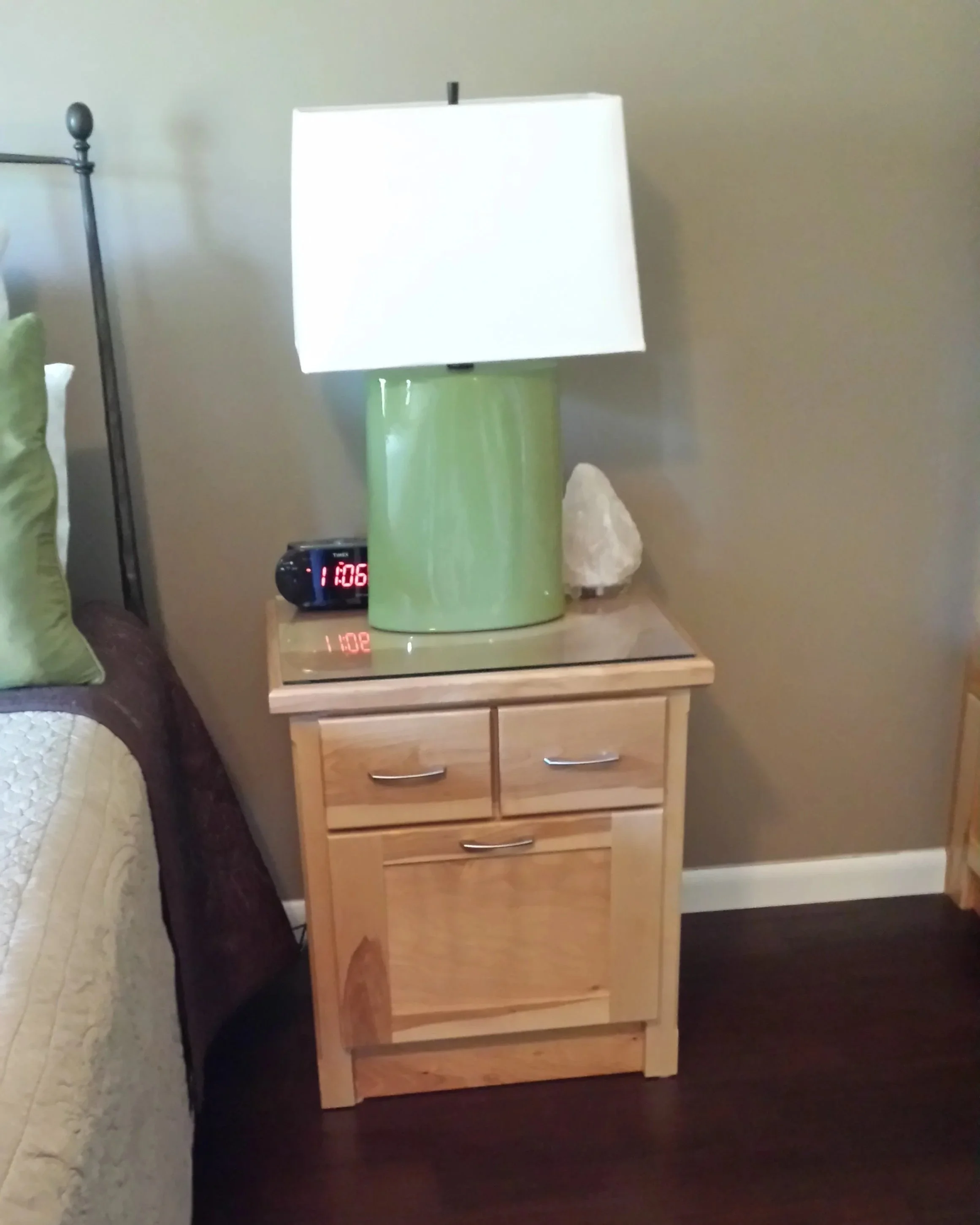 A bedroom nightstand with a green lamp, a digital alarm clock showing 11:06, and a white rock, next to a bed with a green pillow.