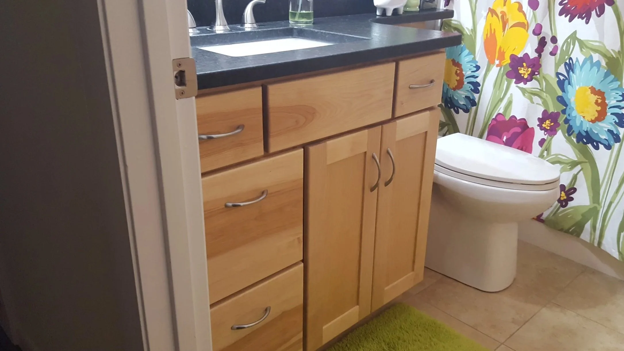 A bathroom with a wooden cabinet, a black countertop, and a white toilet. There is a floral shower curtain with large, colorful flowers and a green bath mat on the beige tiled floor.