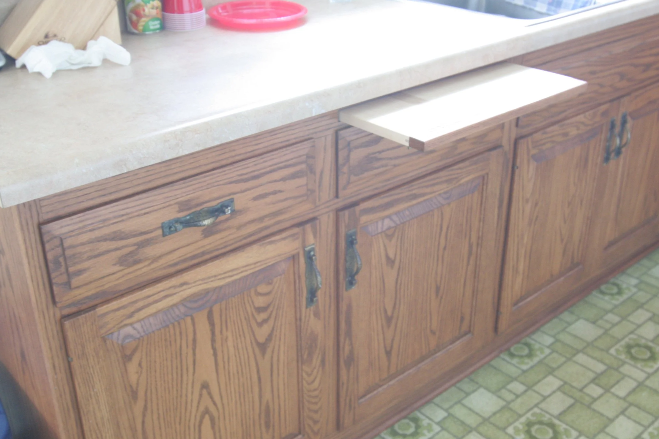 Kitchen cabinet with a open drawer and wooden doors, countertop with some items, and green patterned floor.