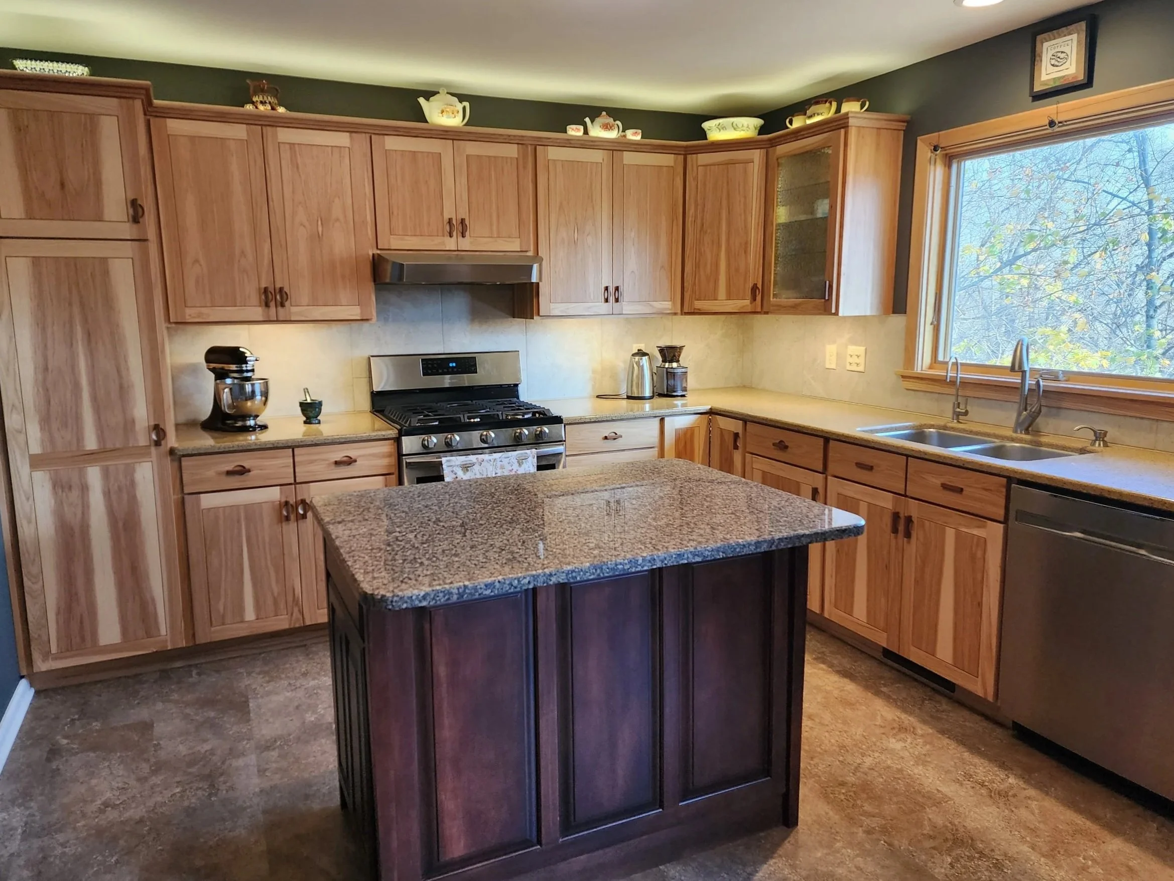 Kitchen with wooden cabinets, granite countertop island, stainless steel stove, double sink, window showing trees outside, and various small appliances on the counters.