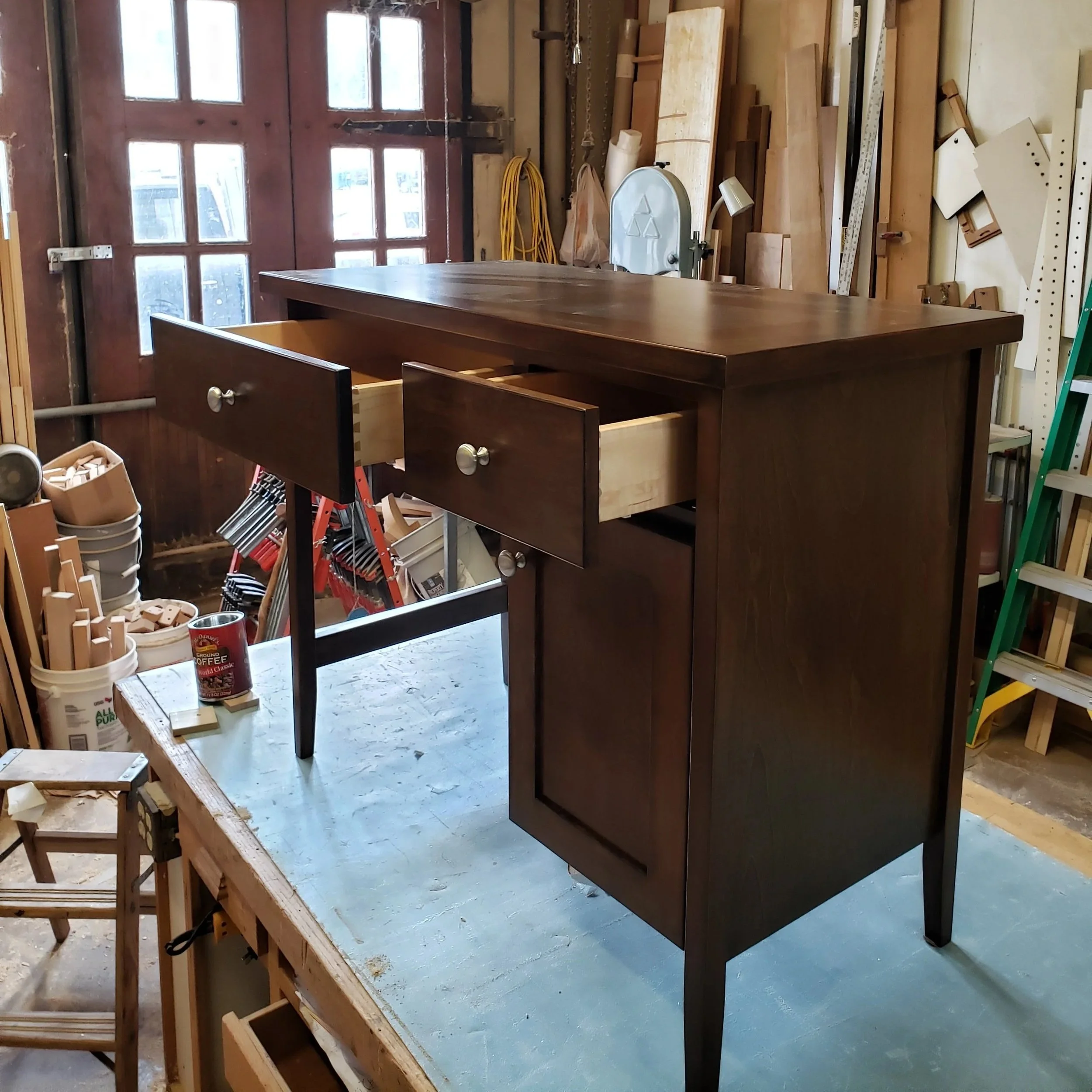 A wooden desk with open drawers is in a woodworking workshop. The workshop has various tools, wood pieces, and equipment in the background.