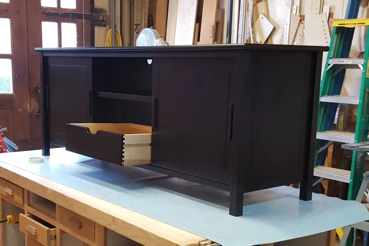 Black wooden sideboard with sliding doors and a partially open drawer, placed on a workbench in a woodworking workshop.