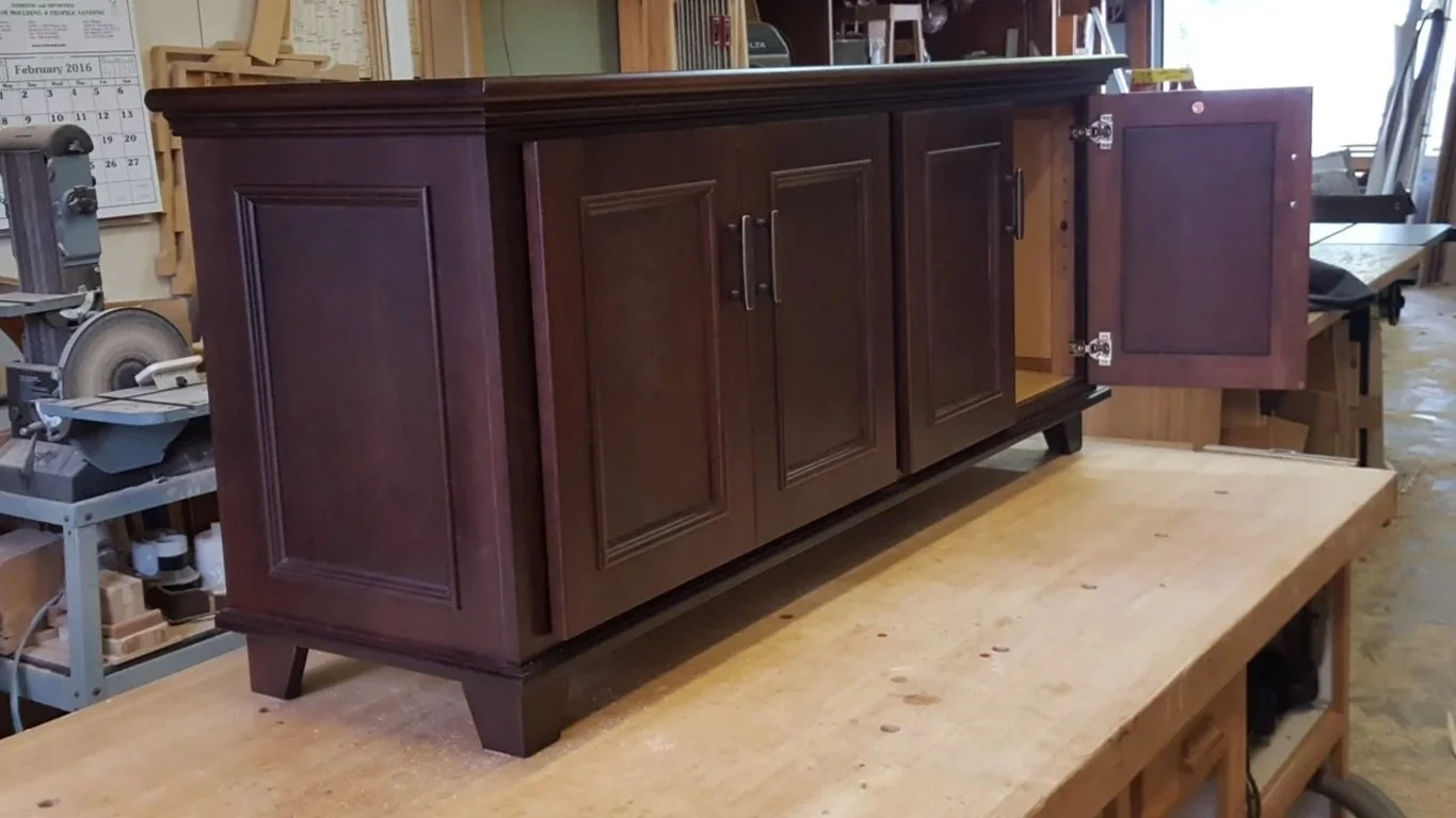 A dark brown wooden cabinet with four doors, two of which are open, in a woodworking shop.