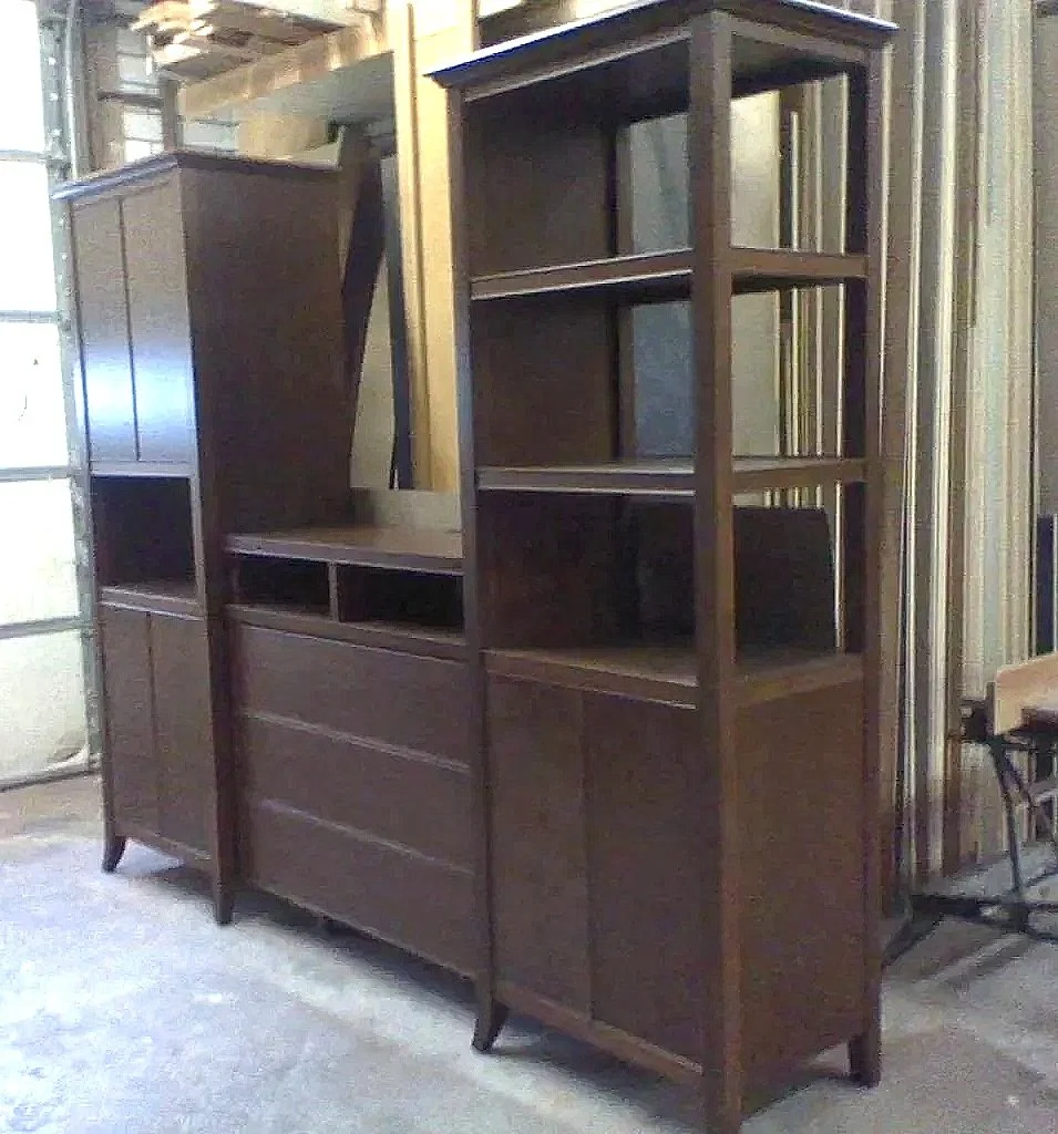 Three tall wooden bookshelves or cabinets with an open section at the top, situated in a workshop or garage with metal siding. The furniture appears to be partially assembled or in storage.