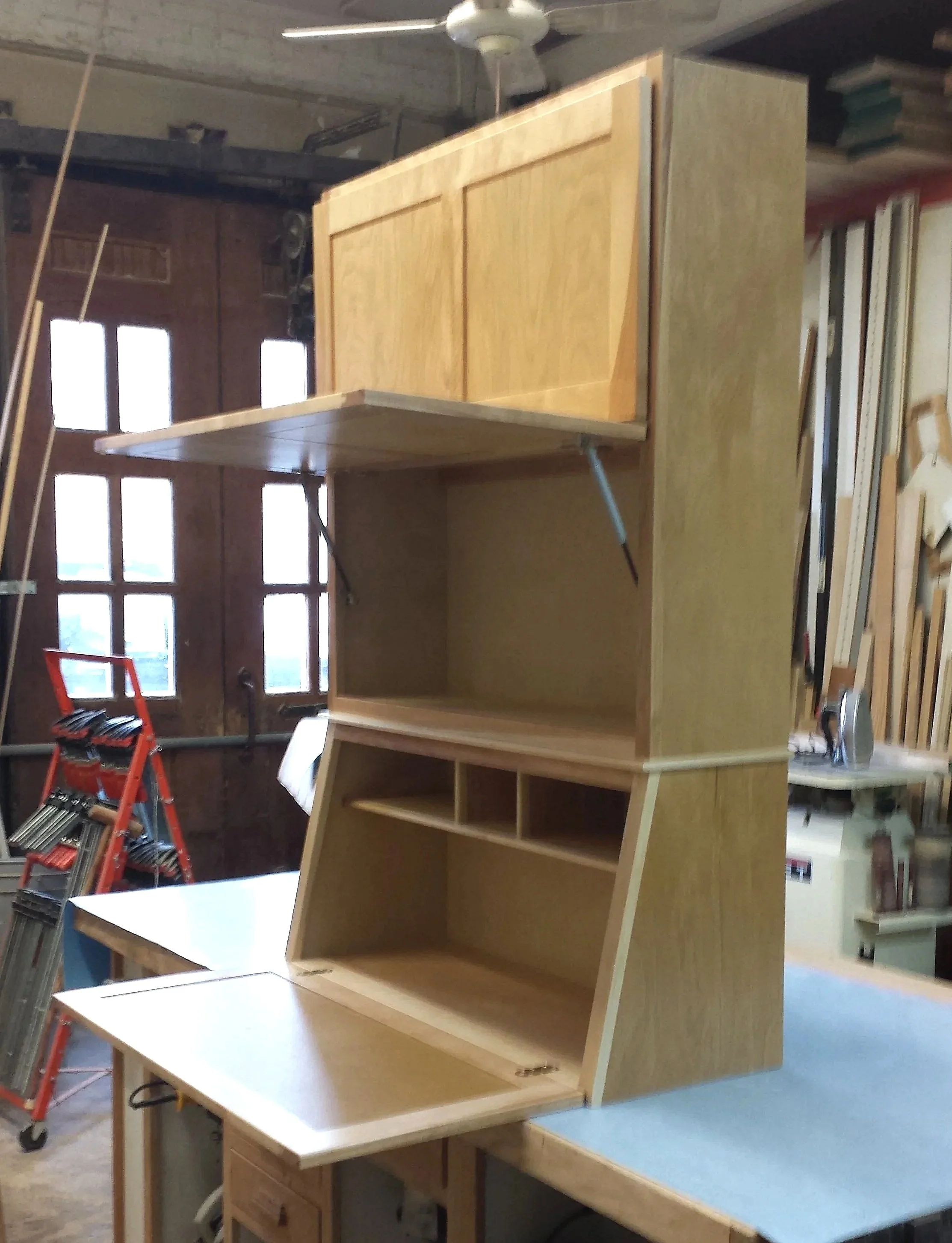unfinished wooden cabinet with multiple compartments and a fold-down door in a workshop.