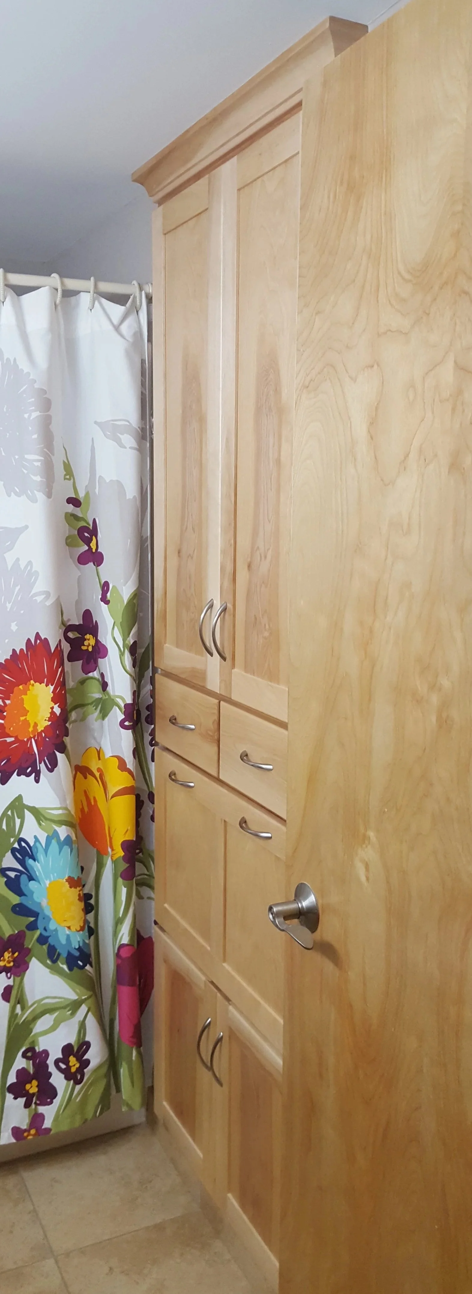 Close-up of a tall wooden bathroom cabinet with silver handles next to a floral shower curtain.