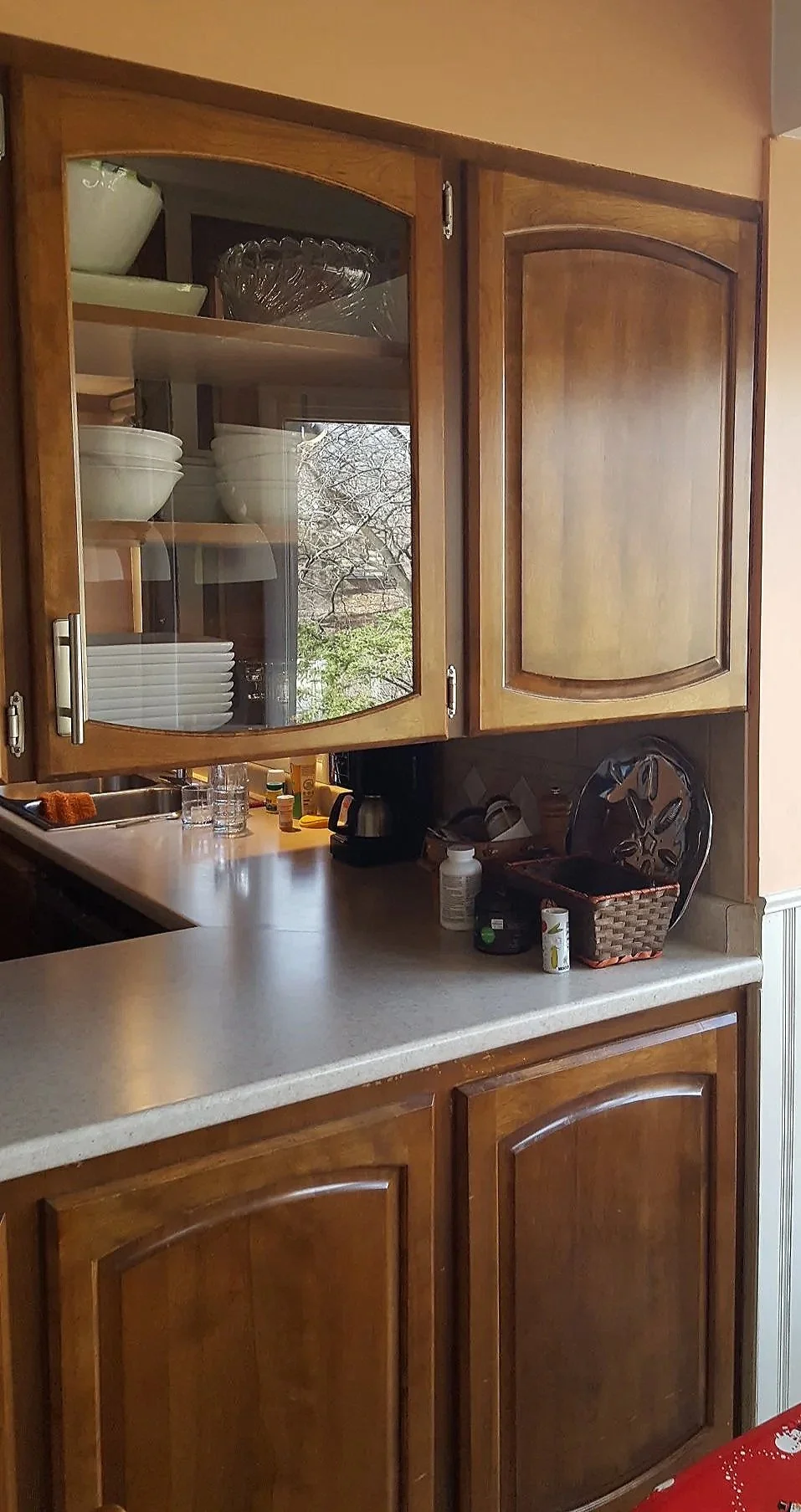 Kitchen cabinets with glass doors displaying bowls and glasses, countertop with bottles, a basket, and some small items, above a lower cabinet with extended countertop.