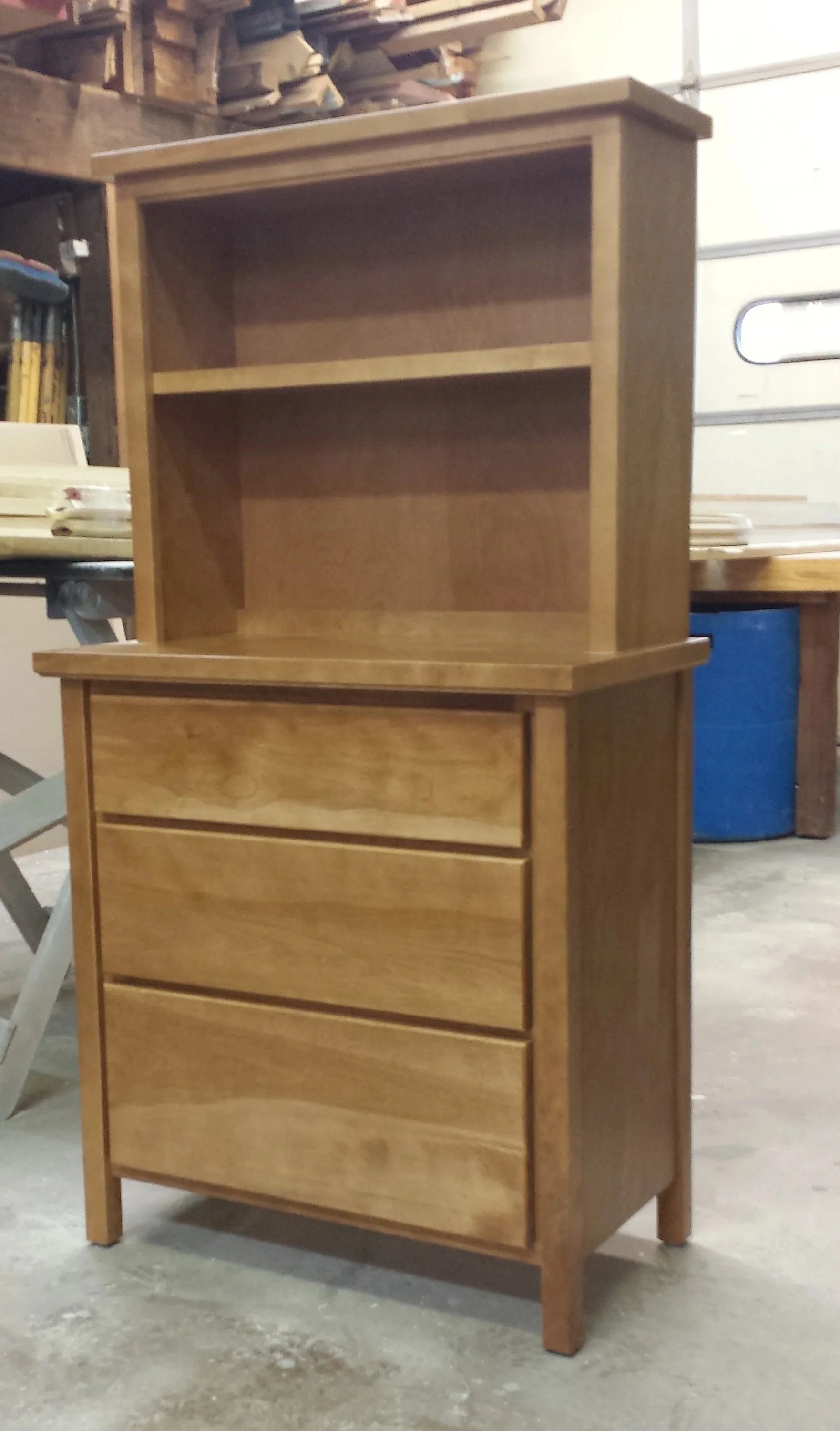 A two-part wooden cabinet with shelves on the top section and three drawers on the bottom, situated in a workshop or storage room.
