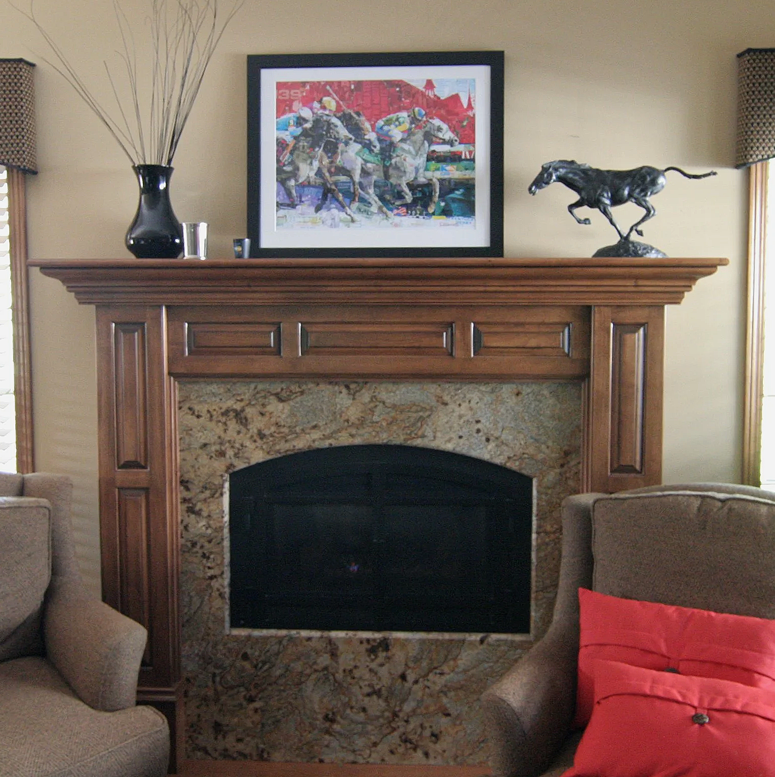 Living room fireplace with wood mantel, picture of horse race, sculpture of running horse, and decorative vase with branches on the mantel, upholstered chairs nearby.
