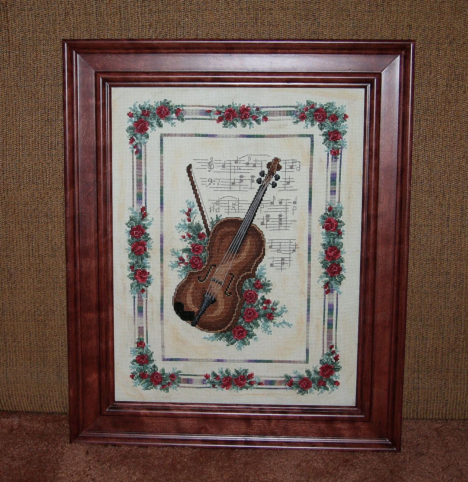 Framed cross-stitch artwork depicting a violin surrounded by red roses and sheet music background.