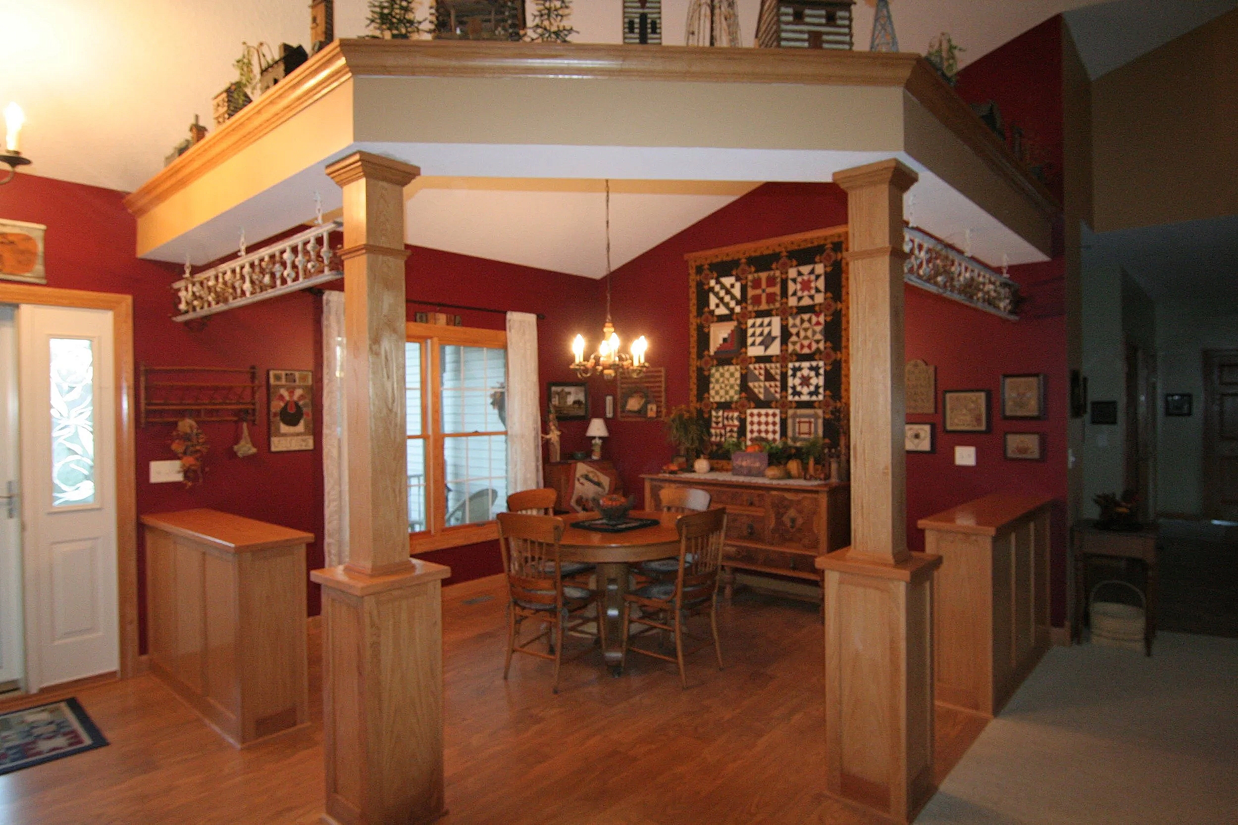 Interior of a dining area with a wooden round table and four chairs, decorated with a chandelier overhead, surrounded by red walls adorned with framed pictures and a quilt, with a sliding door leading outside and various decorative items on shelves a