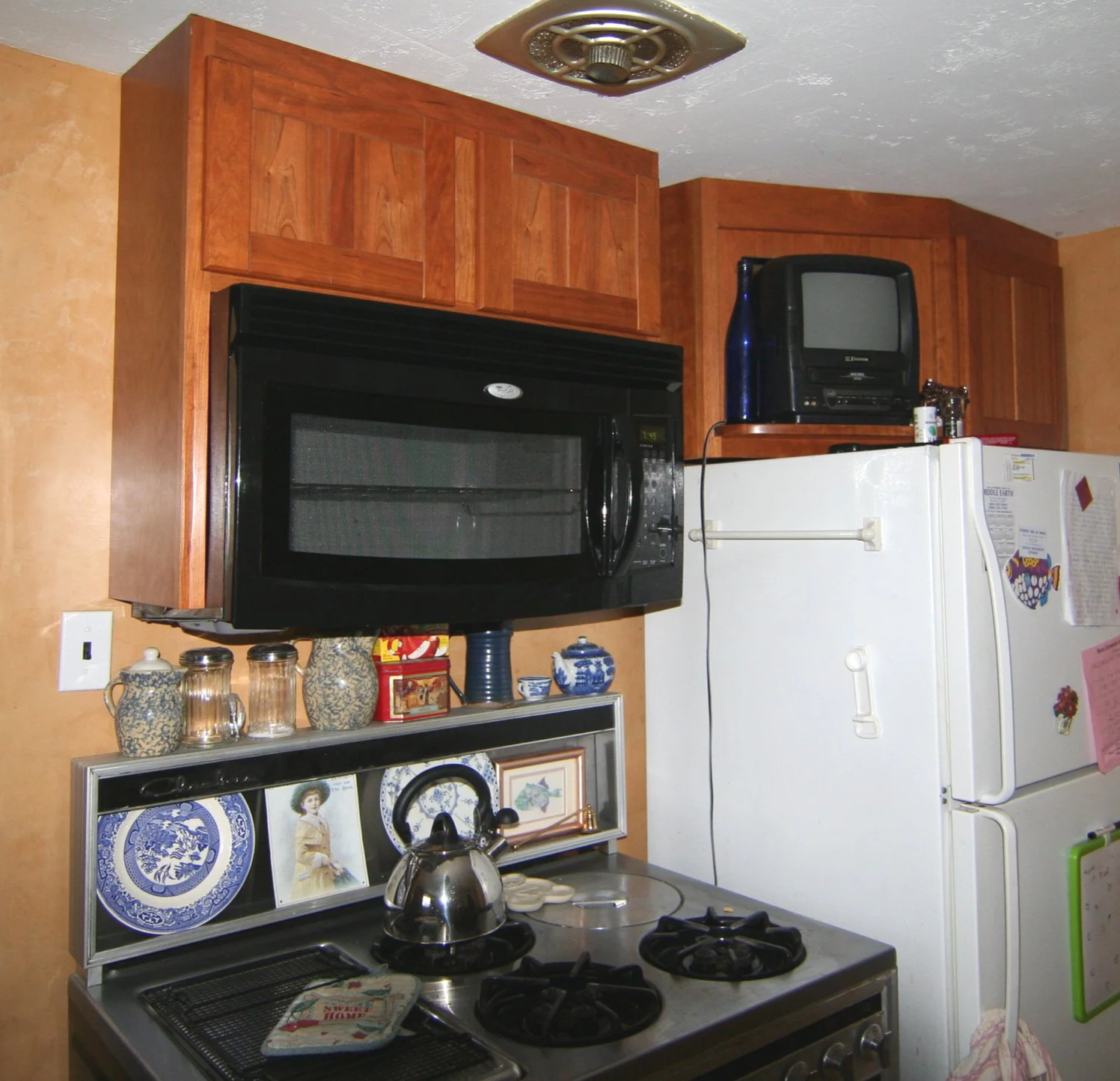 Kitchen with wooden cabinets, black microwave, small old TV on refrigerator, white refrigerator, spice bottles, decorative plates, and a stovetop with a kettle.