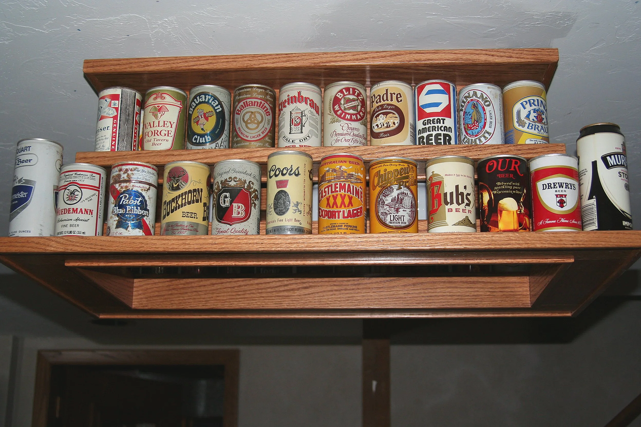 A wooden shelf mounted on a ceiling holding metal cans of various beer brands.