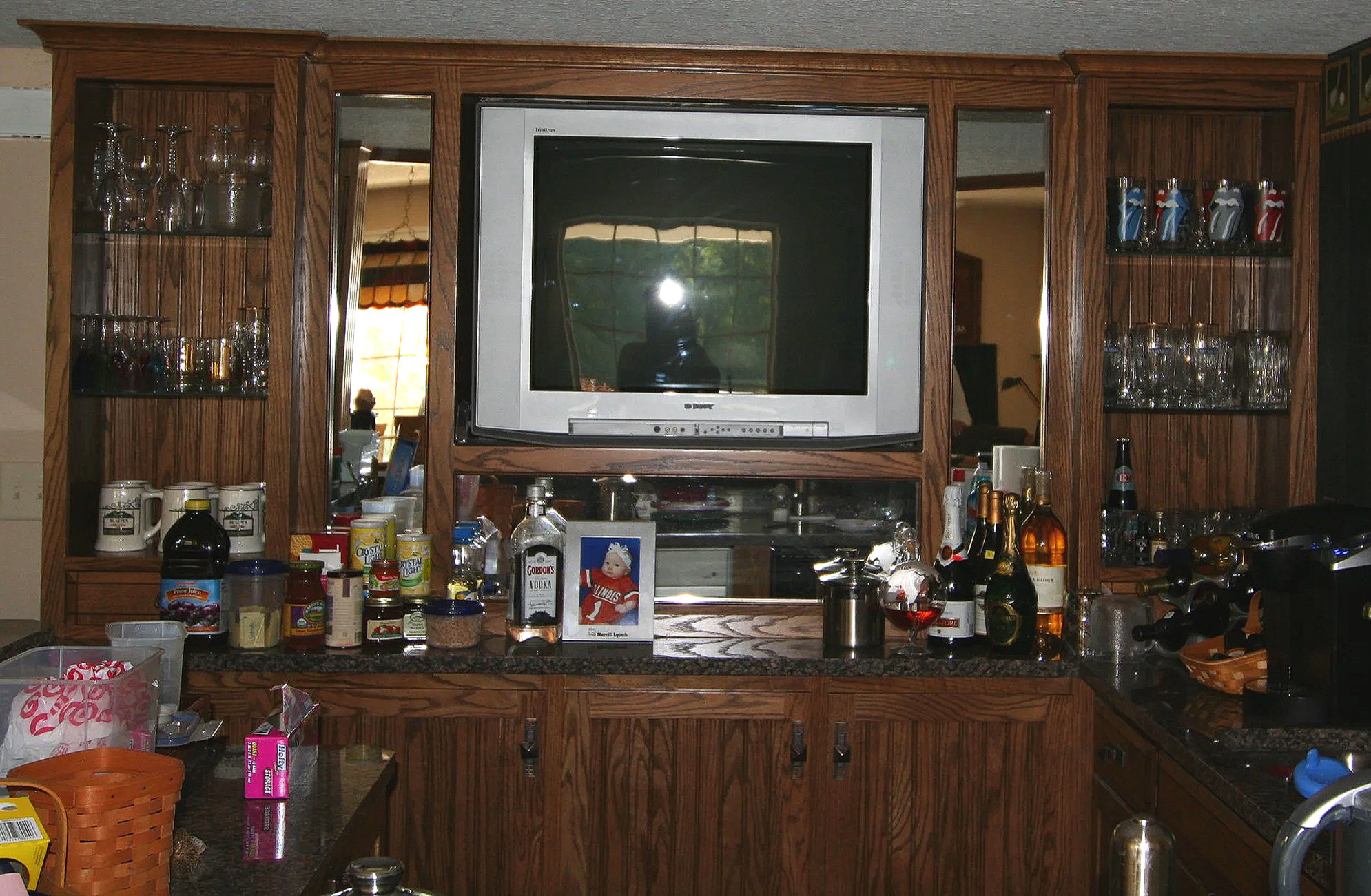 A cluttered kitchen counter with various bottles, cans, and containers, and a mounted TV in a wooden cabinet with glassware and decorative items.