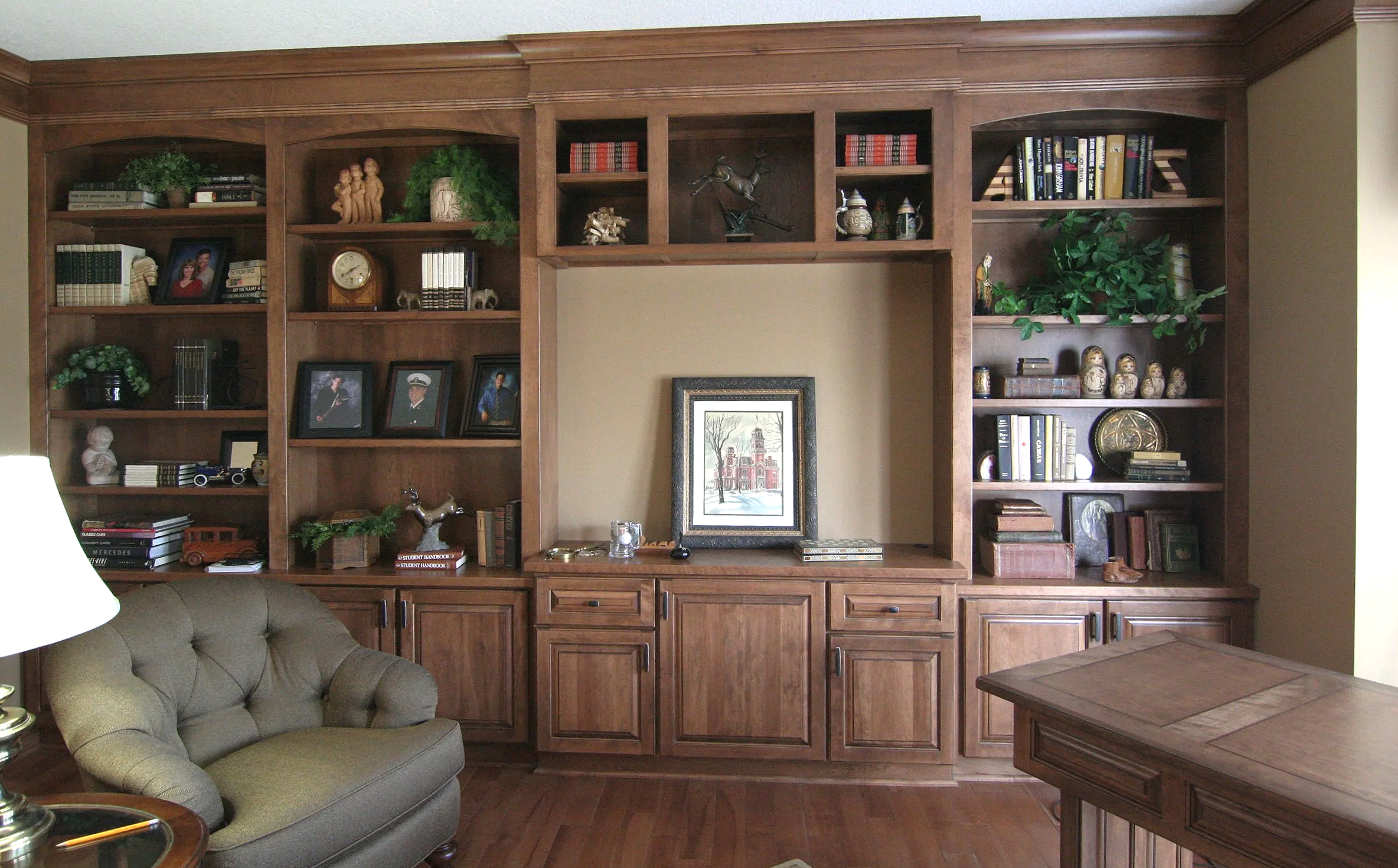 A large wooden built-in bookshelf with various framed photographs, books, decorative items, and plants, with a beige wall background and a wooden floor.