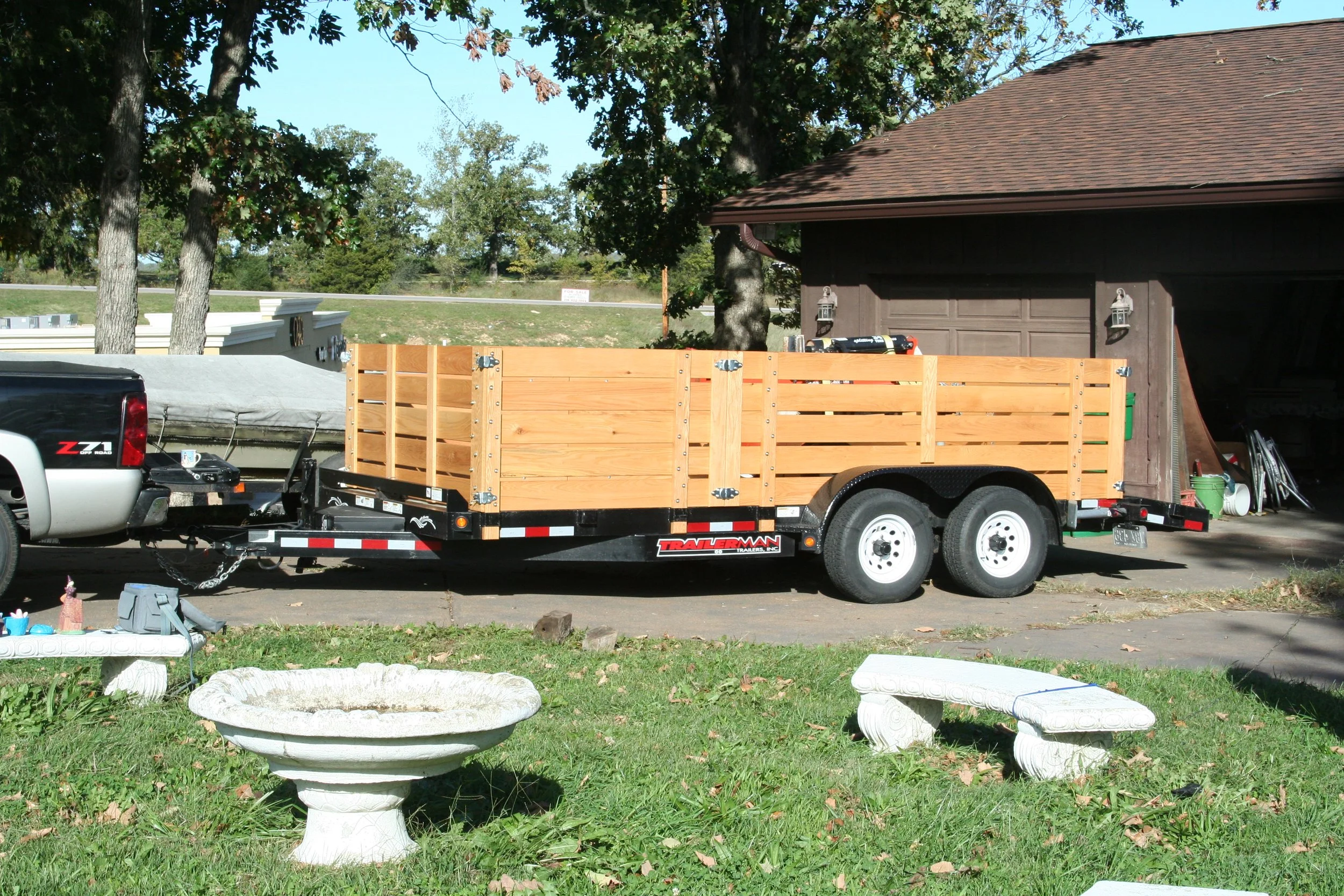 A house with an open garage, a wooden trailer attached to a pickup truck, and outdoor garden furniture in the front yard.