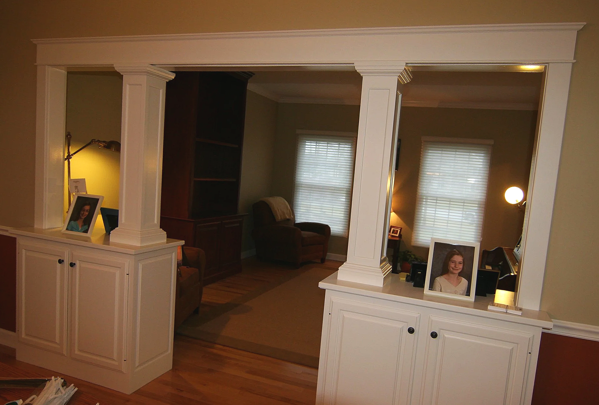  view through a large cutout in a wall showing a living room with hardwood floors, two windows with blinds, a brown armchair, and various lamps and pictures.