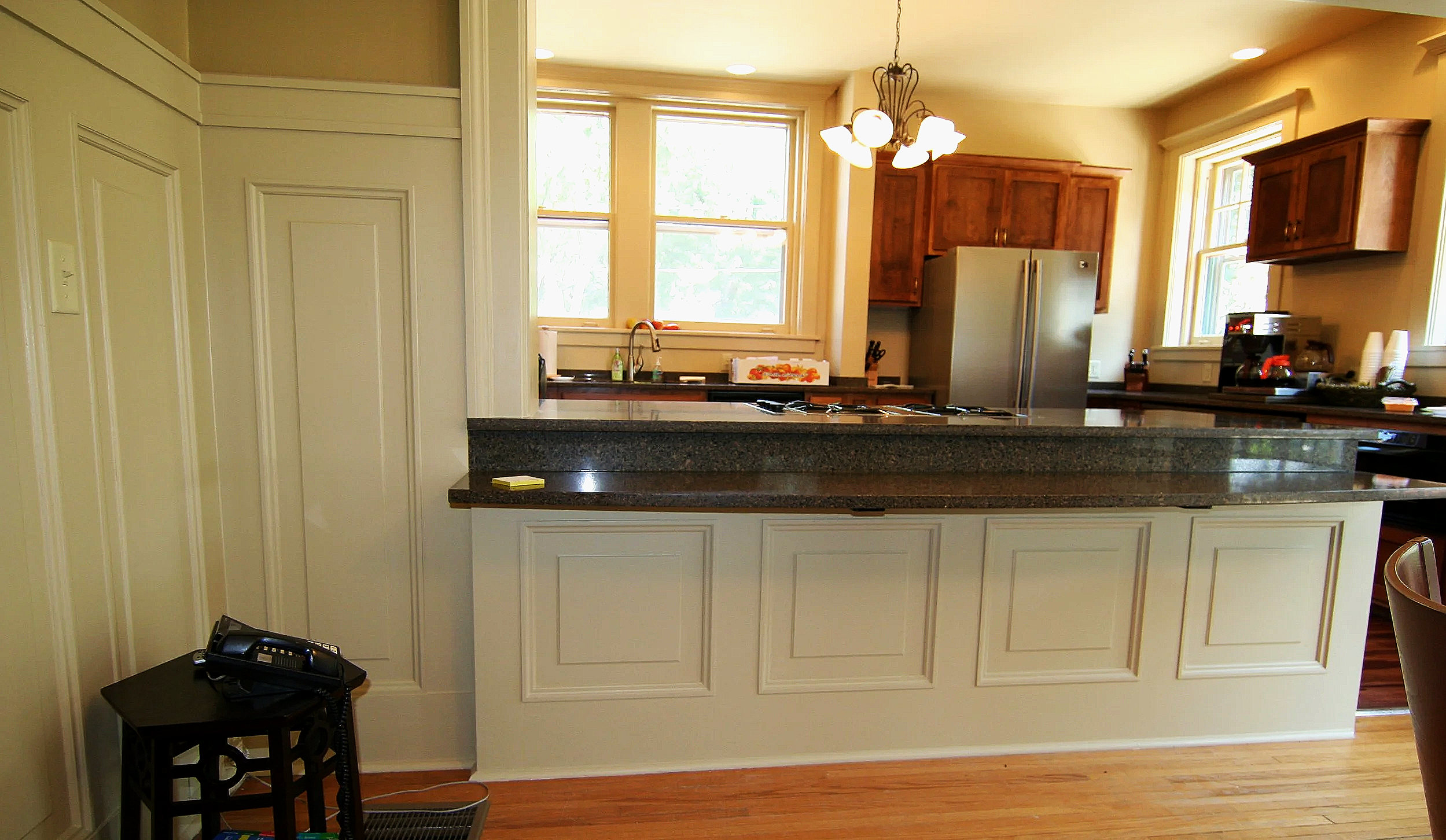 A kitchen with wooden cabinets, a stainless steel refrigerator, a granite countertop, and two windows letting in natural light. There is a chandelier hanging from the ceiling, and various kitchen appliances and items on the countertops.