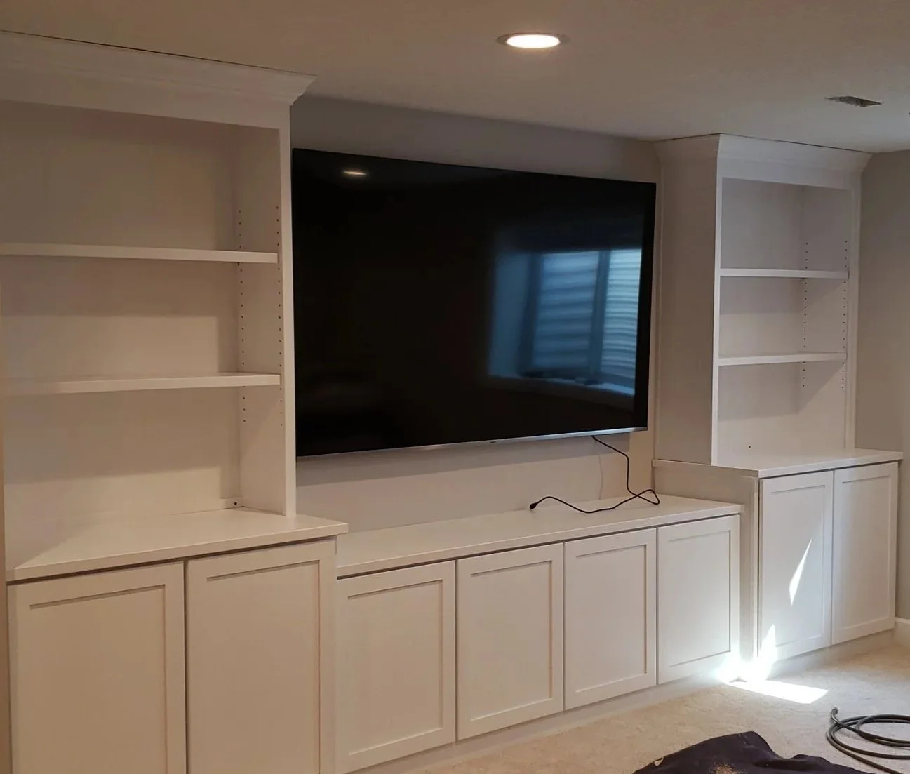Built-in white entertainment center with open shelves and closed cabinets, black flat-screen TV, and a window reflection visible on the TV screen.
