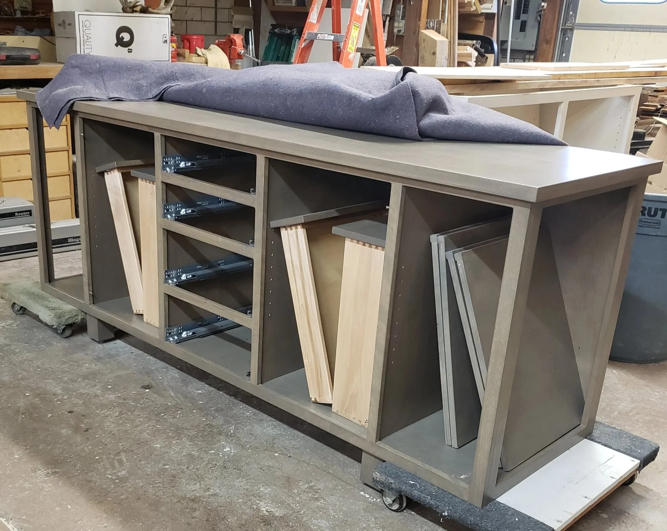 The image shows a partially assembled wooden cabinet or entertainment center in a workshop, with three sections containing metal drawer slides and wooden panels, and a gray cloth draped over the top.
