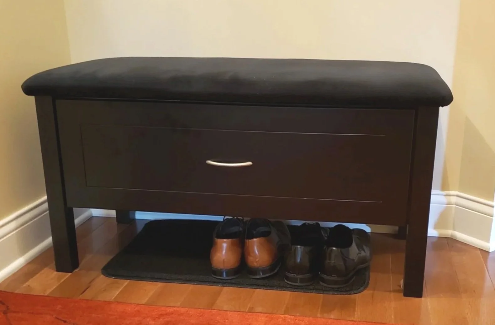 A black wooden shoe storage bench with a cushioned seat on top. There are three pairs of shoes underneath it on a black mat, placed against a beige wall on a hardwood floor.